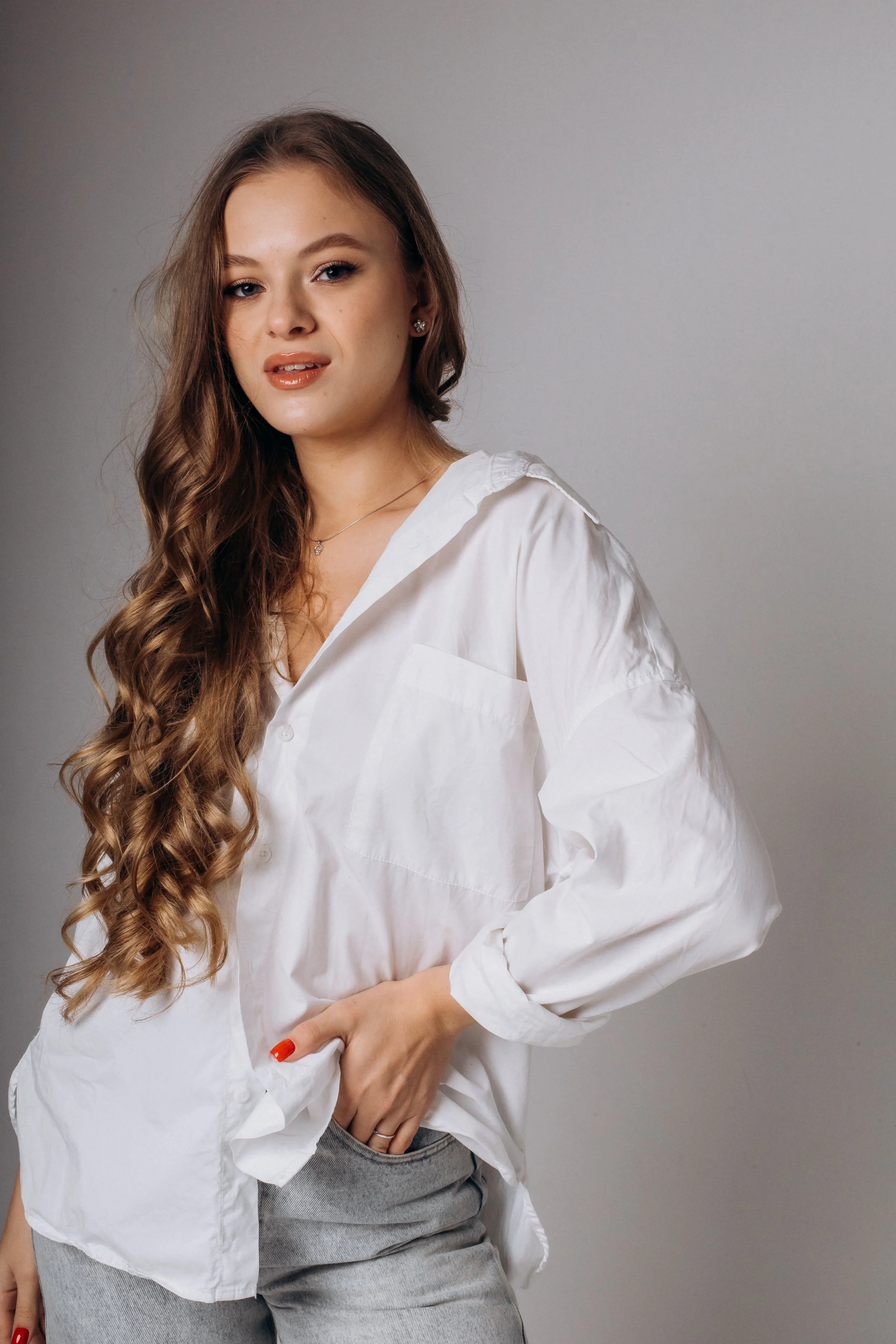 A young woman with long, wavy brown hair, wearing a white button-up shirt and gray pants, posing against a neutral background.