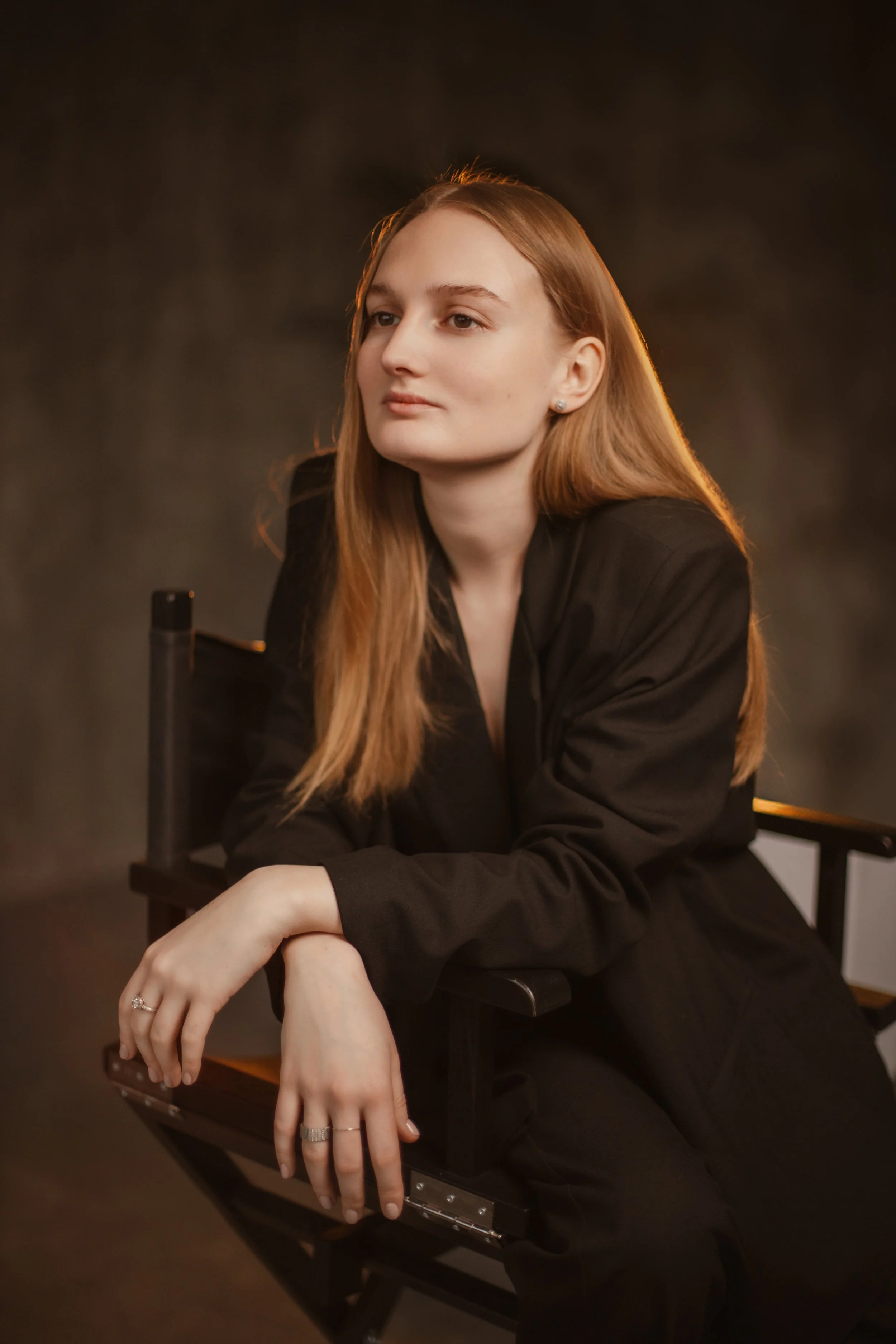 Portrait of a young woman with long red hair, wearing a black jacket, sitting on a director's chair against a dark background, looking to the side.