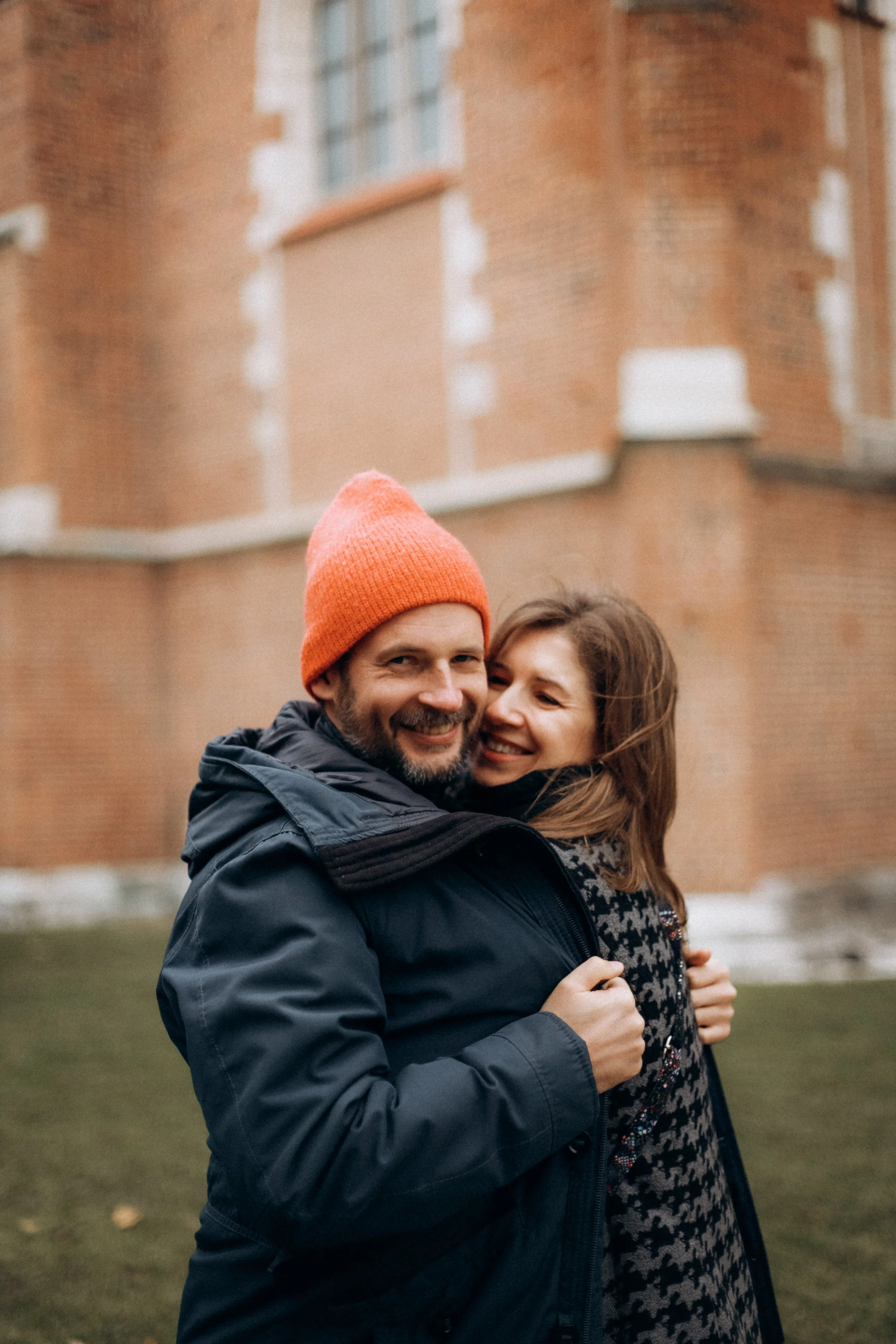 A man and woman smiling and hugging outdoors in front of a brick building during fall or winter.