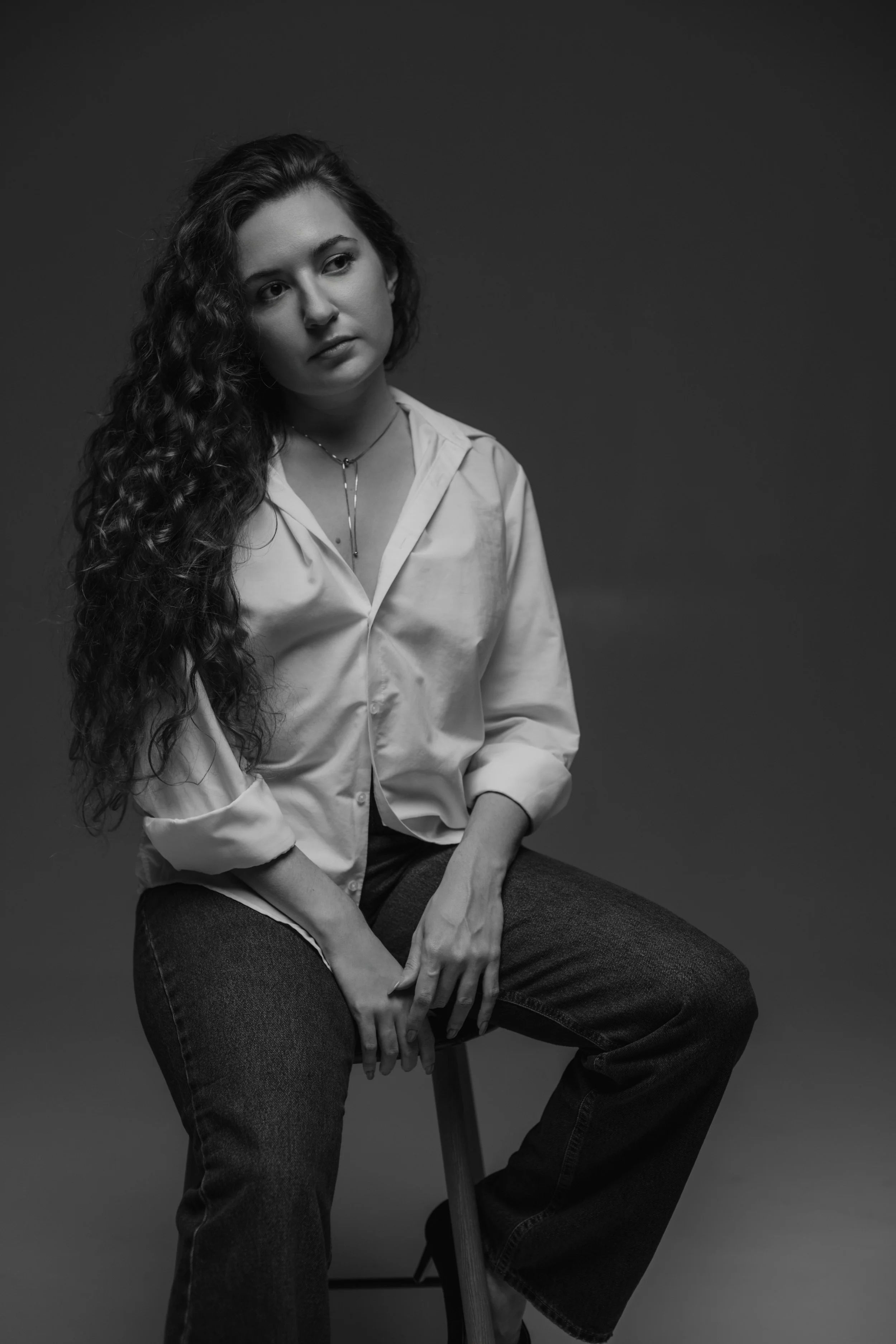 Black and white portrait of a young woman with long, curly hair, sitting on a stool against a dark background, wearing a loose shirt and jeans.