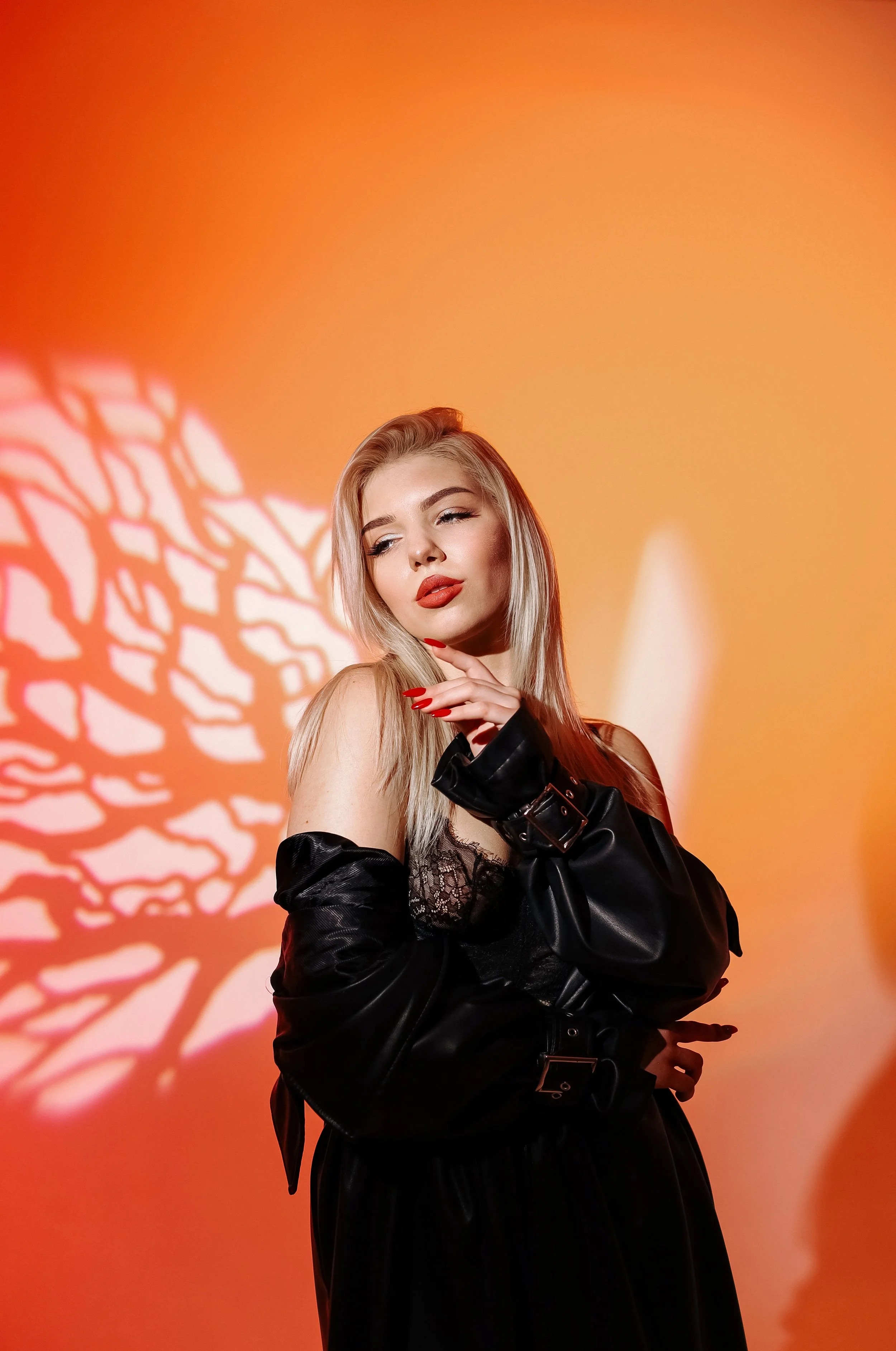 A young woman with blonde hair and red lipstick posing with her hand near her chin against a warm, orange background with a shadow of a leafless tree.