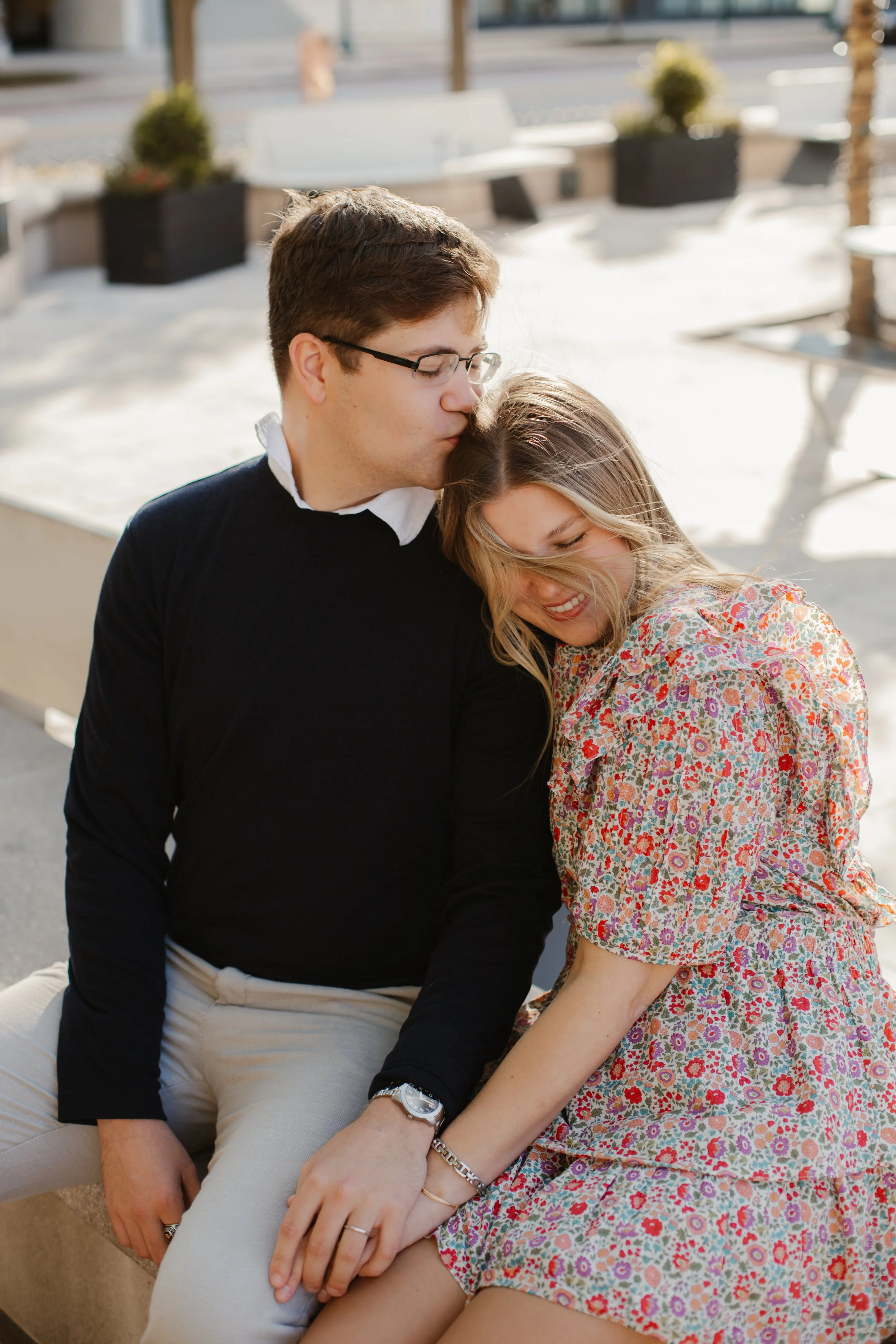 A man and woman sitting close together outdoors, holding hands, with the man kissing the woman's head, both smiling and appearing happy.