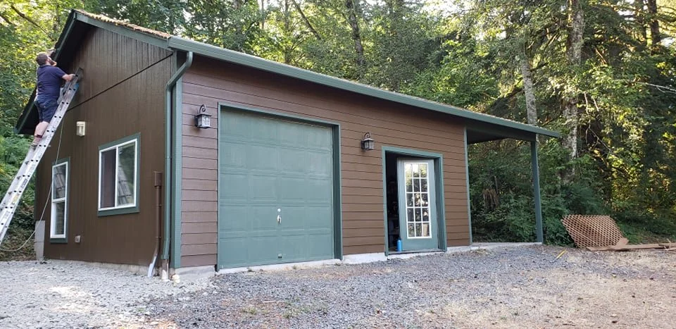 A person on a ladder painting the exterior of a small brown garage or shed with a large green door and a glass-paneled door, situated in a wooded area.