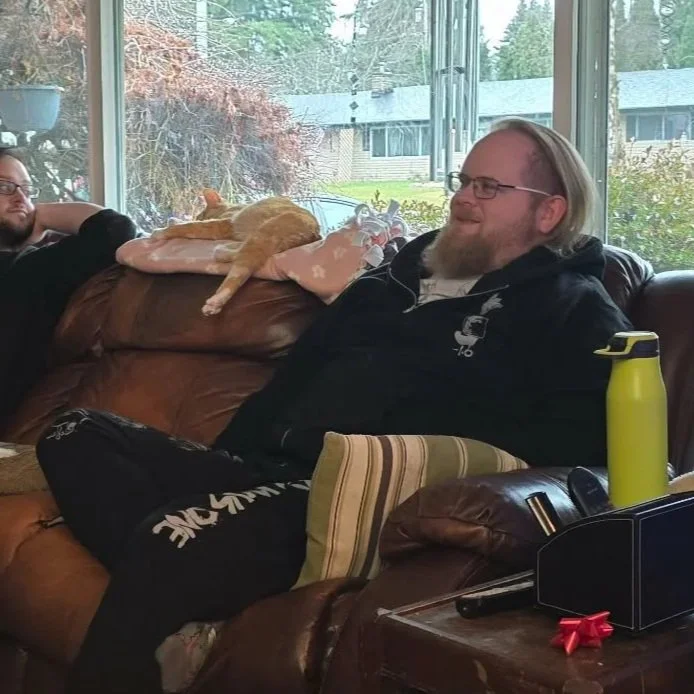 A man with a beard and glasses sitting on a brown leather couch, smiling. On the couch, an orange cat is sleeping on a pillow near his shoulder. A second person is partially visible on the left. A yellow water bottle is on a side table next to him. Behind, large windows show trees and a house outside.