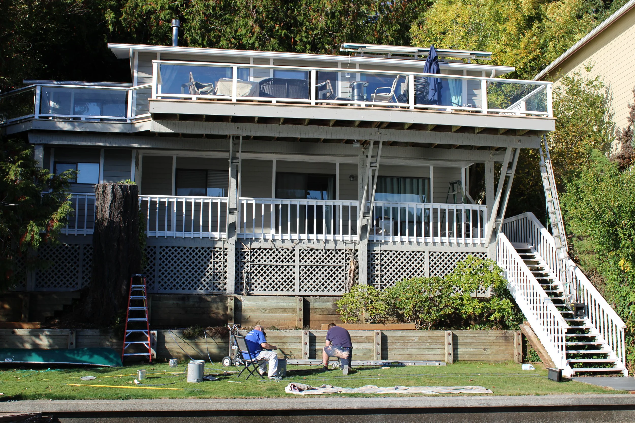 Two workers working on a construction project on the lower level of a house with a large deck and staircase, surrounded by trees and shrubs.