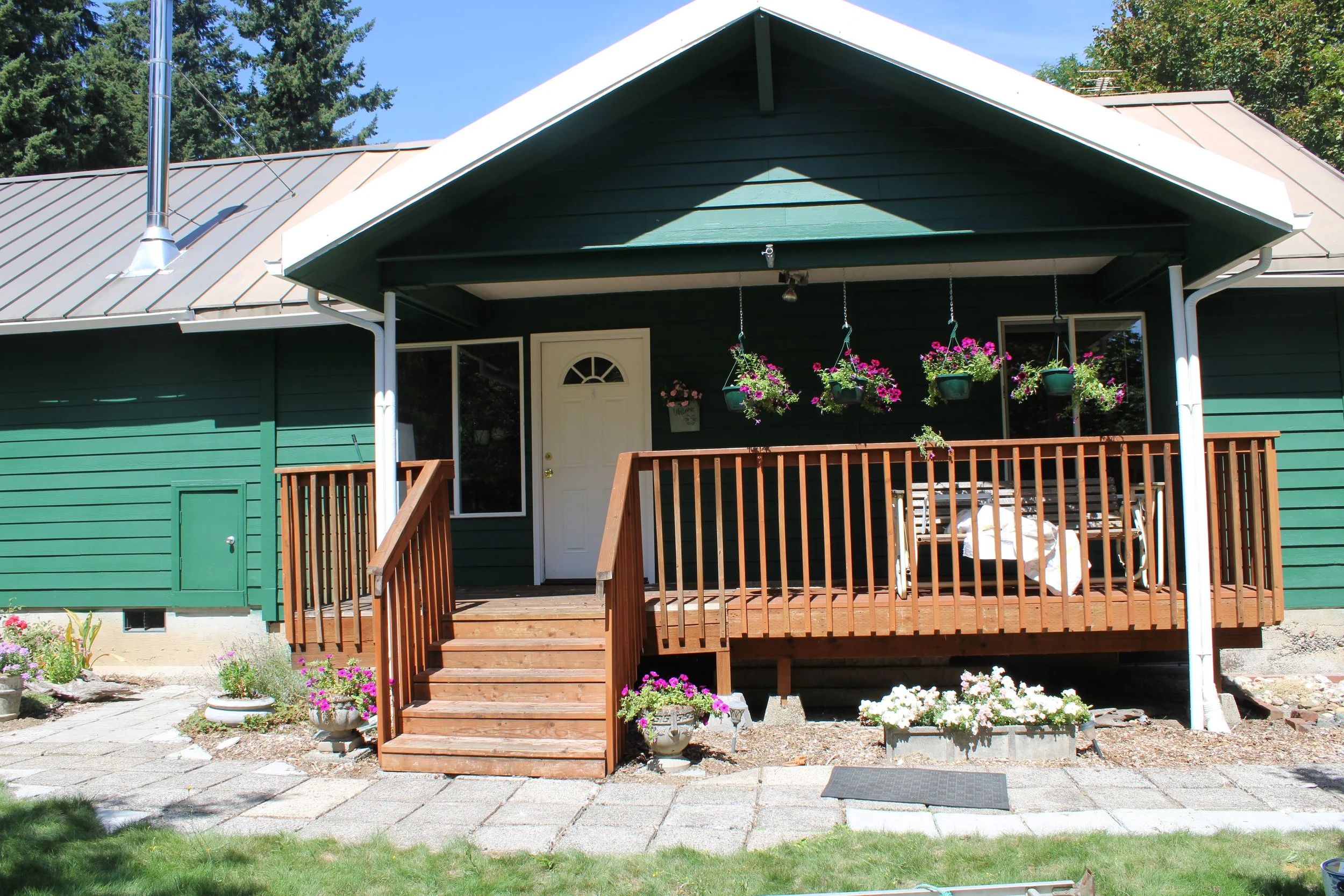 Front view of a green house with a porch, wooden stairs, hanging pink and purple flowers, and potted plants, with a stone pathway and lawn in front.