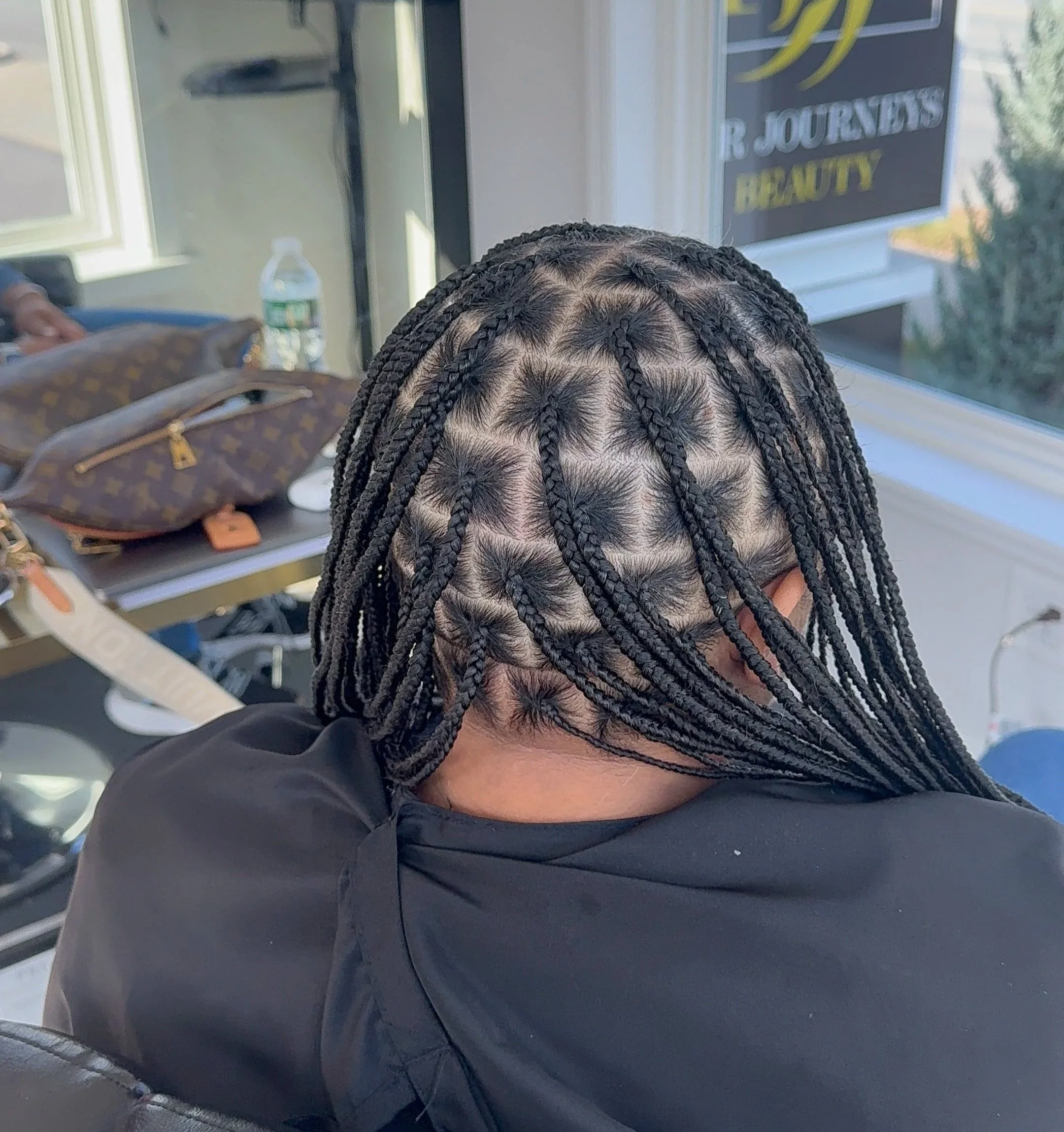 A person with freshly done box braids hairstyle, sitting in what appears to be a salon with a window, some bags, and a water bottle on the surface behind them.