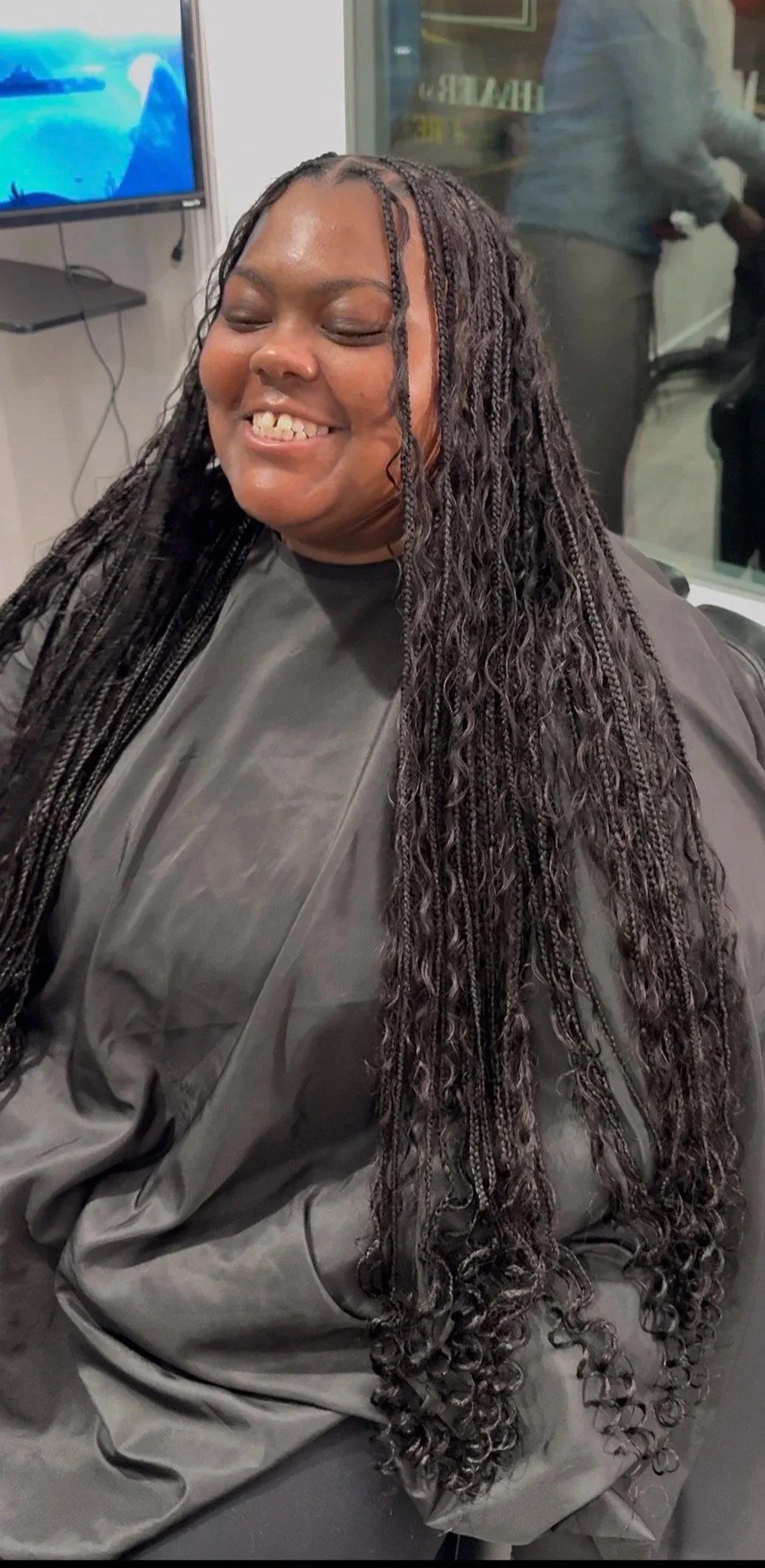Woman with long, curly braids smiling with eyes closed, seated at a salon with a black cape around her shoulders, mirror and TV screen in the background.