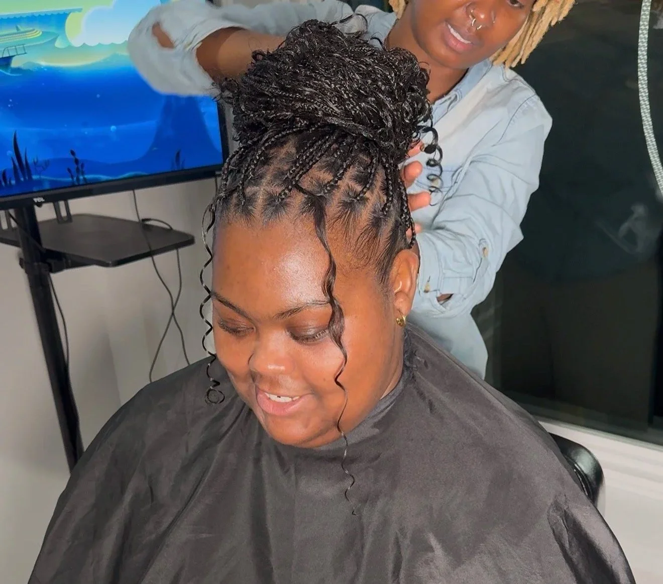 A woman getting braided hairstyle in a salon, with a stylist working on her hair, a man in the background, and a television screen displaying an animated scene.