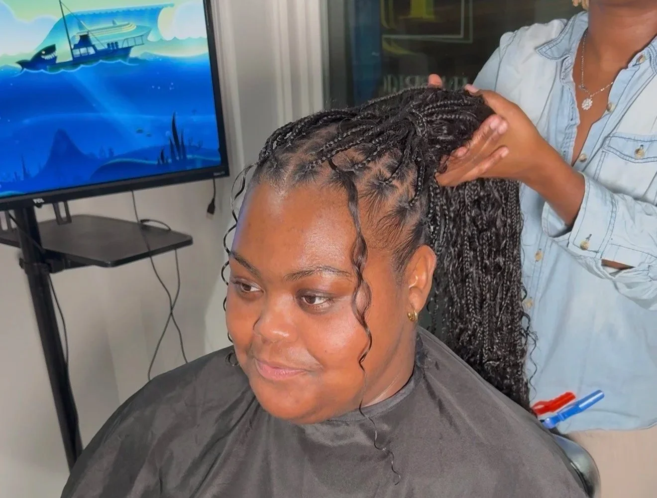 A woman getting a hairstyle with small braids, while seated in a salon chair. A hairstylist is seen holding her braided hair, working on her hairstyle. A television screen in the background displays a cartoon image of a boat sailing on water.