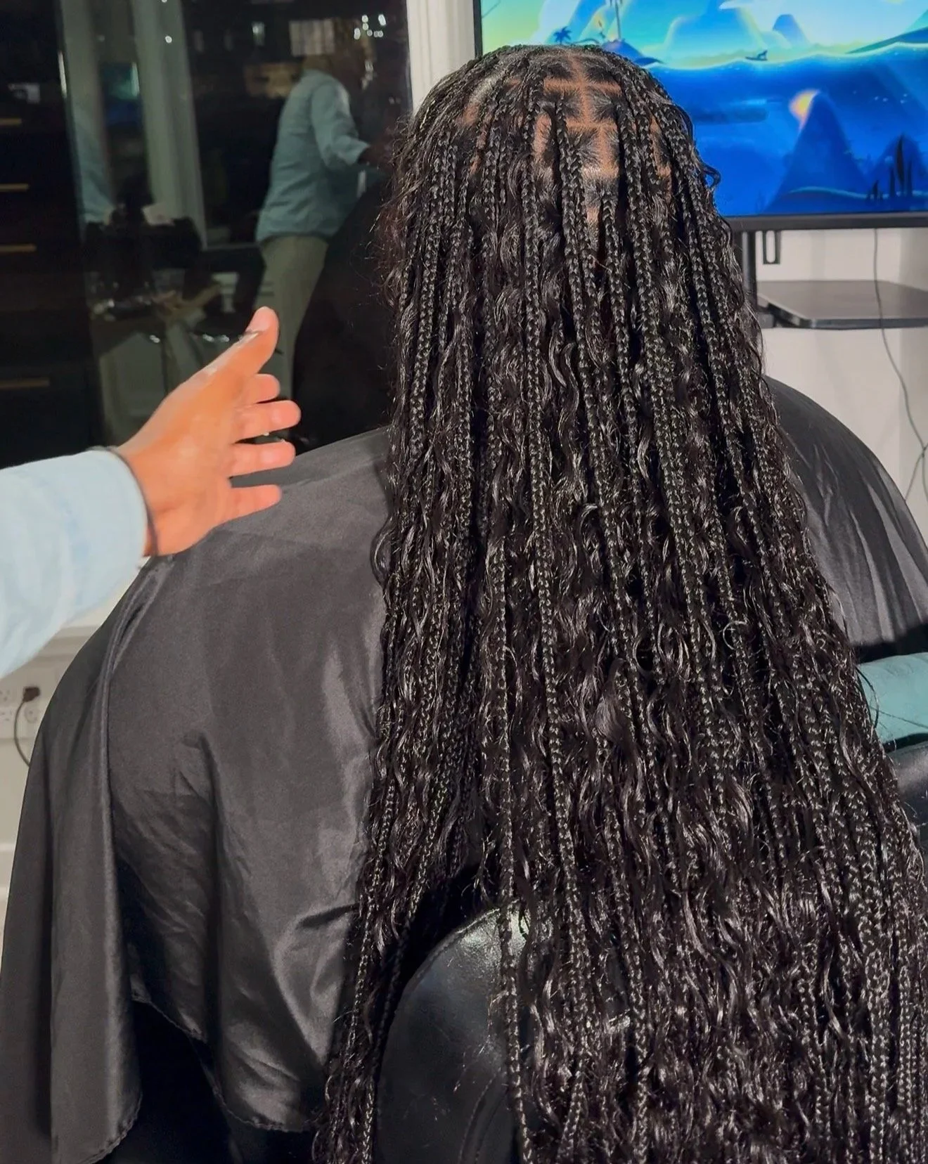 A woman with long, braided, curly hair sitting in a salon chair, with her back facing the camera, and a stylist's hand reaching nearby.