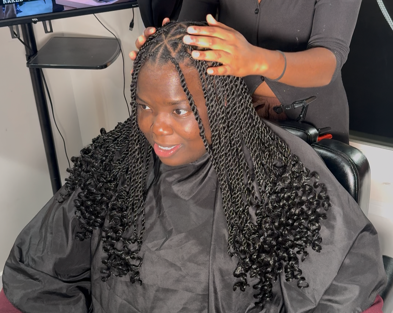 A woman with long, dark, twisted braids is sitting in a salon chair, getting her hair styled by a stylist who is standing behind her, hand on her head. The woman is smiling, wearing a black salon cape, and the stylist is dressed in a dark gray shirt.