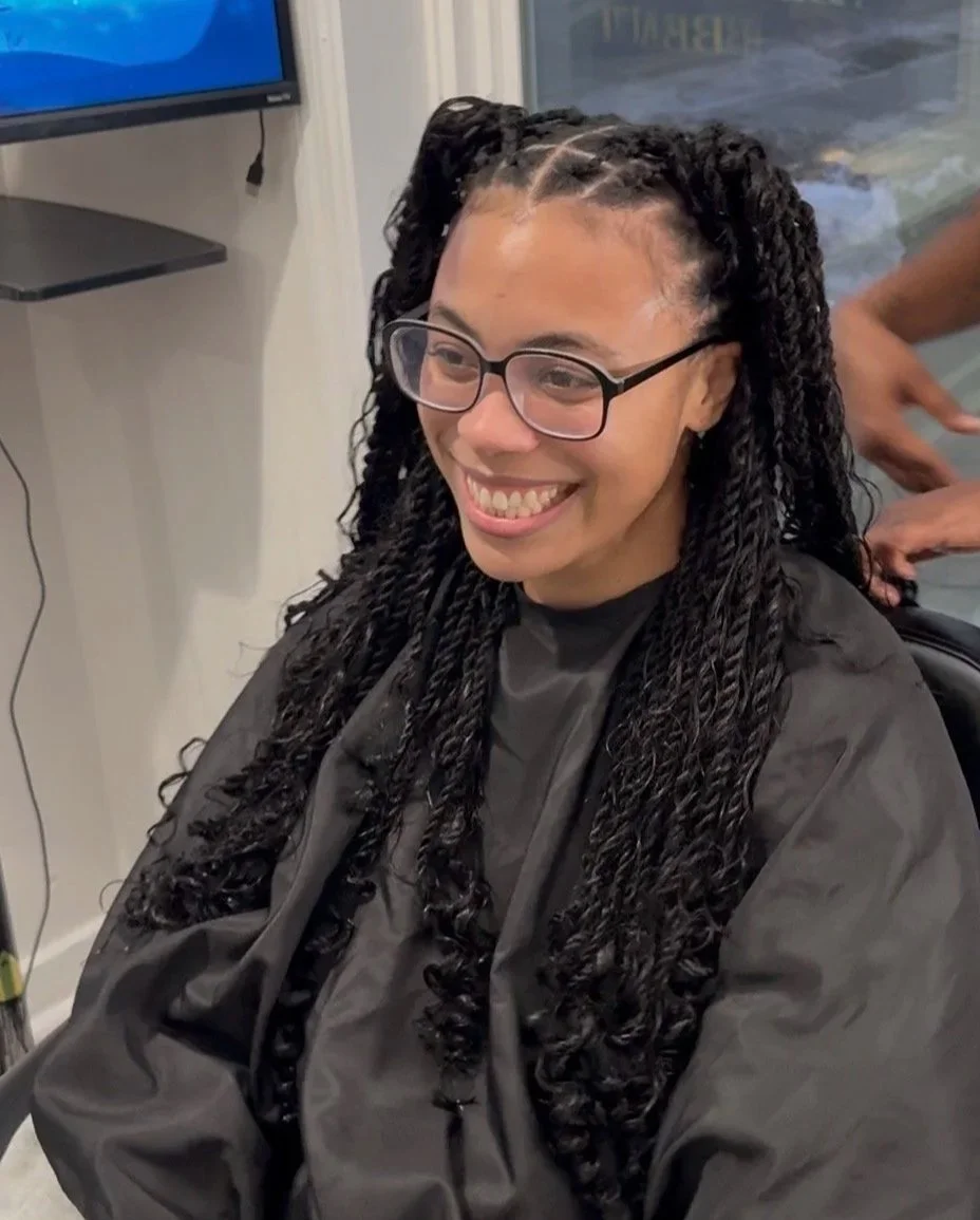 A woman with long, twisted braids and glasses, smiling while sitting in a salon chair wearing a black cape.