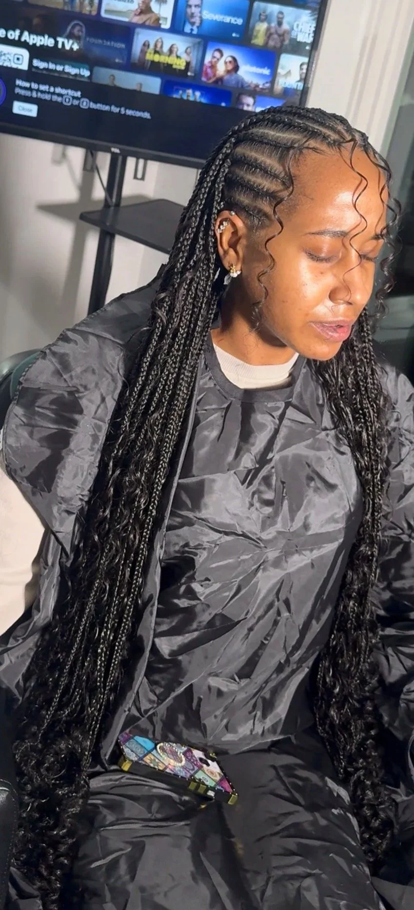 A woman with long braided hair, wearing a black smock, sitting in front of a TV screen displaying streaming service logos, holding a phone with a colorful patterned case, with her eyes closed.