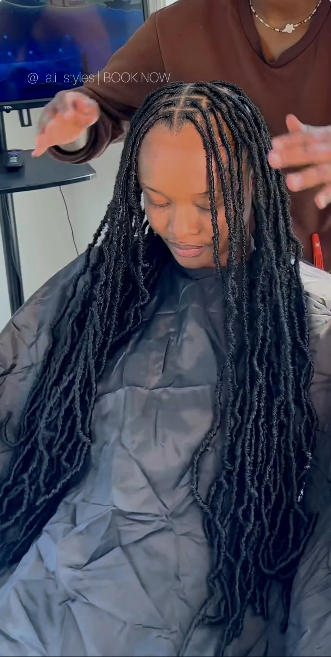 Woman with long black braids seated in a salon chair, receiving a hair treatment or styling. A person standing behind her is working on her hair.