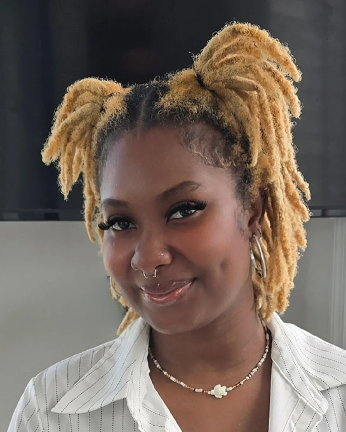 A woman with blonde dreadlocks styled in two high puffs, smiling with jewelry and a white striped shirt.