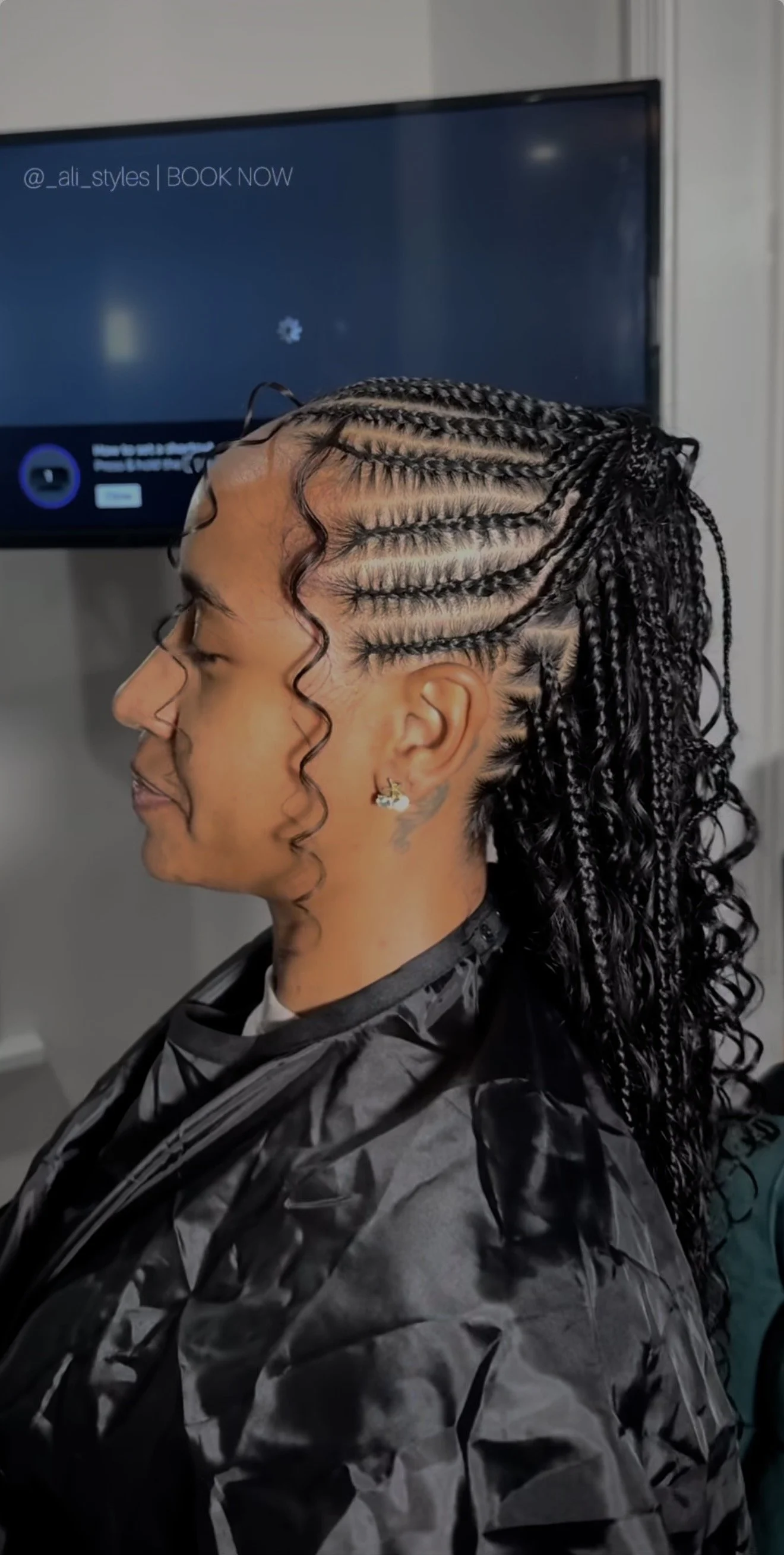 A woman with detailed braided and curly hairstyle sitting in a salon chair, wearing a black cape, with a monitor in the background.