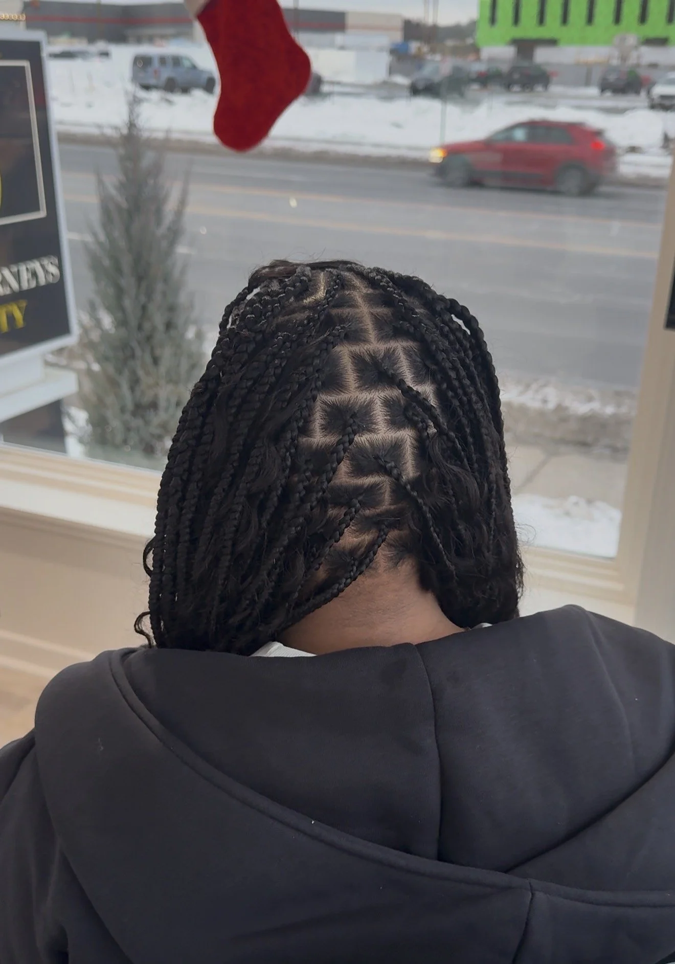Person with braided hair, sitting with their back to the camera near a window showing a snowy street and moving cars outside. A red stocking hangs from the ceiling.