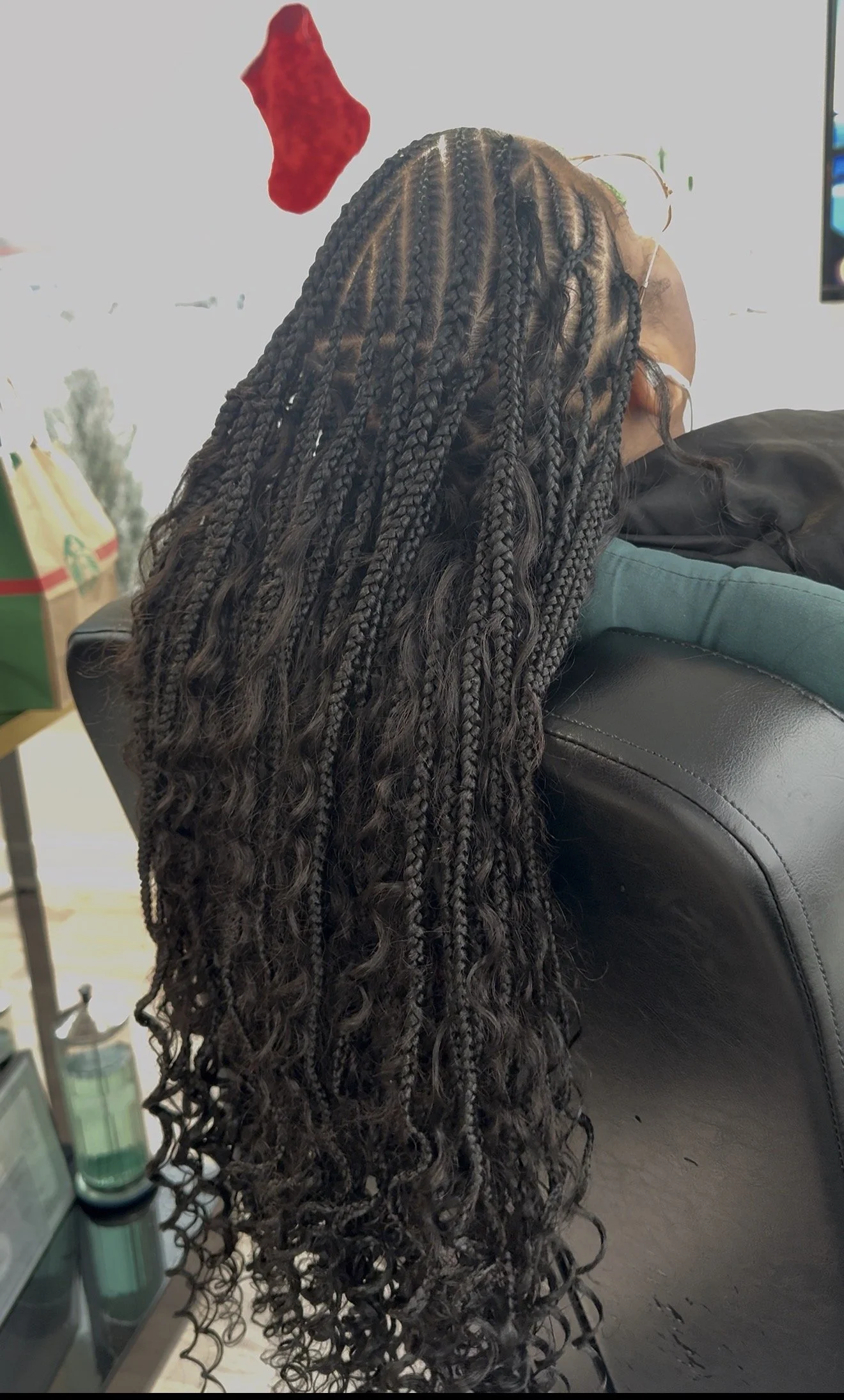 Person with long, black, twisted braids and curly hair sitting on a black leather couch, wearing glasses and a face mask.