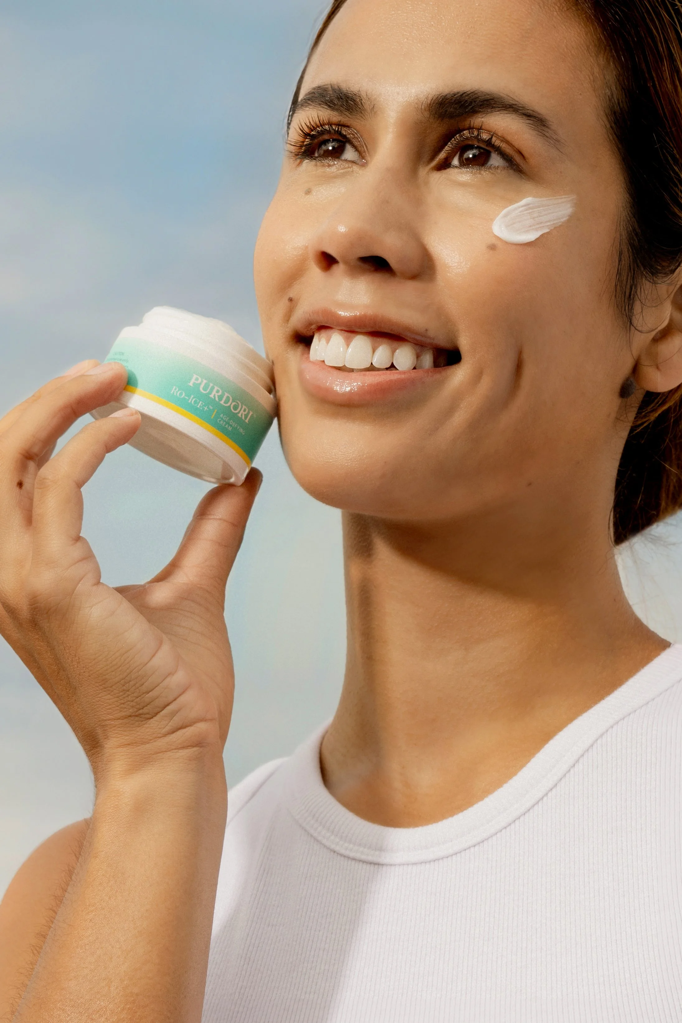 A woman smiling outdoors with sunscreen on her cheek, holding a container of PURDORO skin moisturizer.