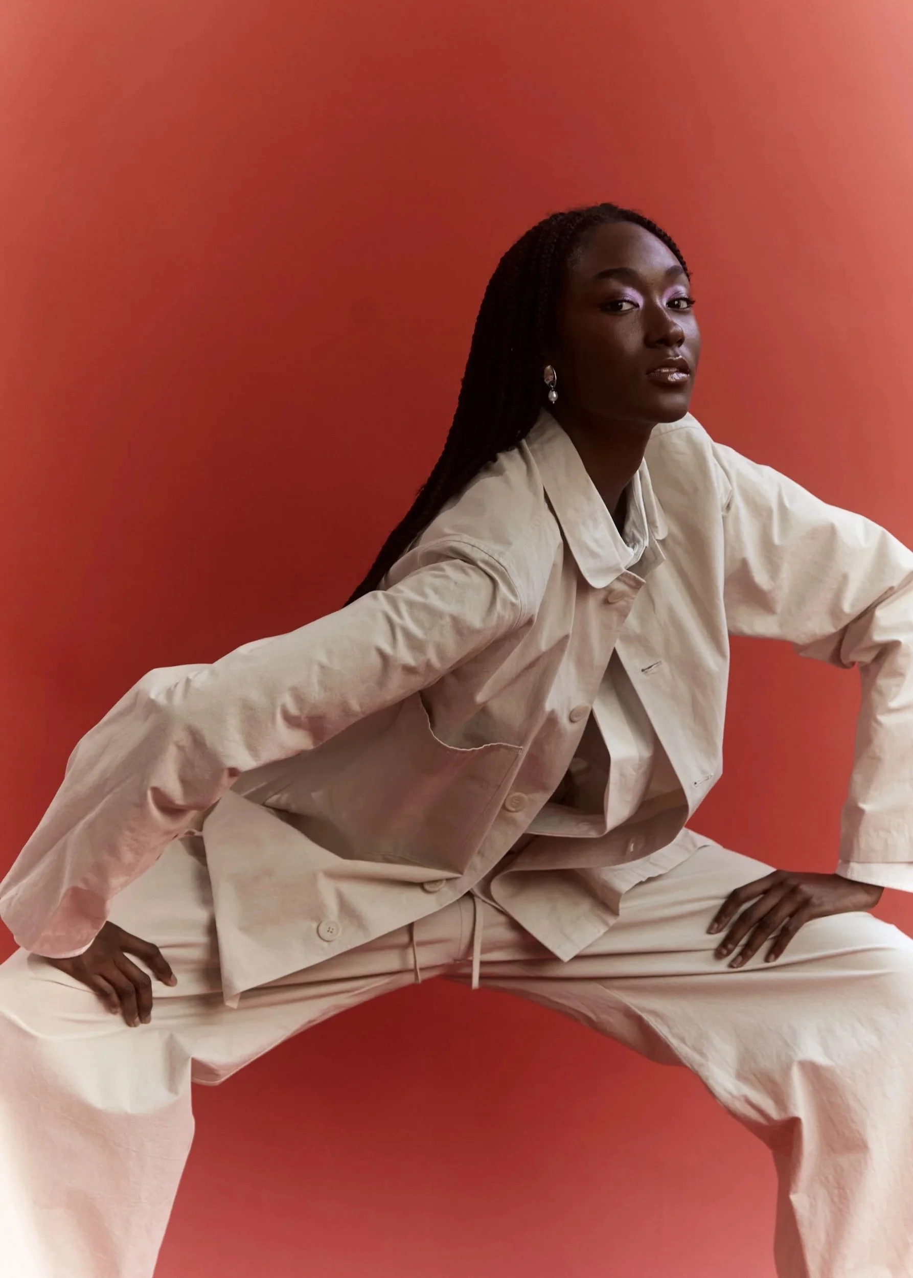 A woman with braided hair wearing a beige oversized jacket and beige pants, posing with her hands on her thighs against a solid coral background.