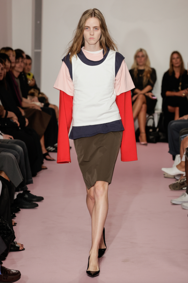 Fashion model walking on runway at a fashion show, wearing a layered outfit with a mix of colors, an olive skirt, and black heels, with an audience seated on both sides.