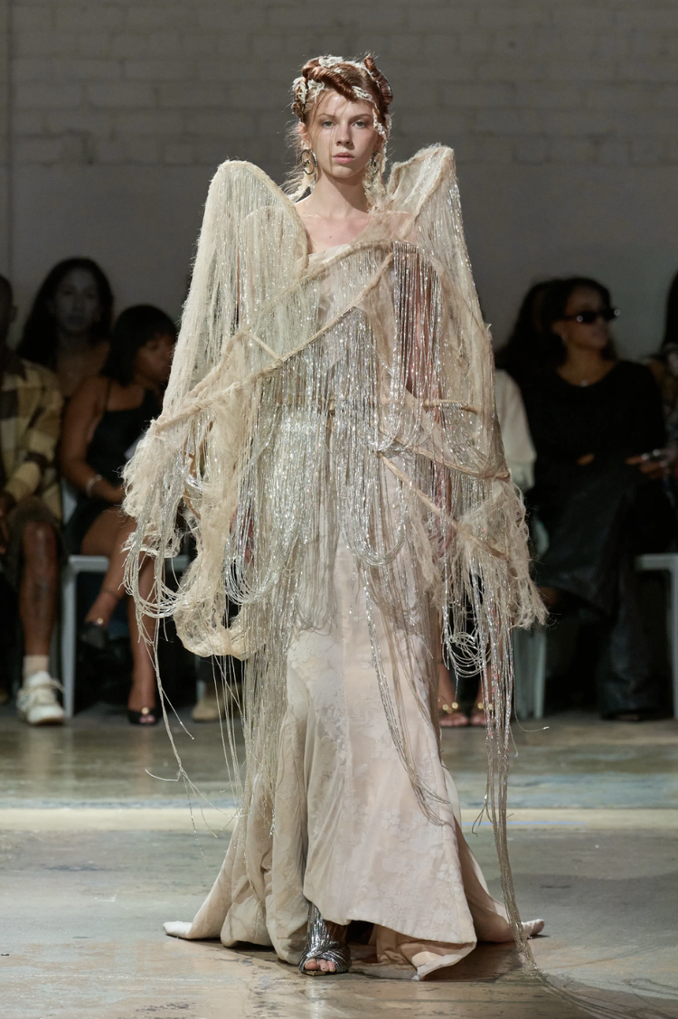 A female model walking down a runway, wearing an avant-garde, beige, and gold metallic dress with large, textured shoulders and metallic fringe details, in front of a seated audience.