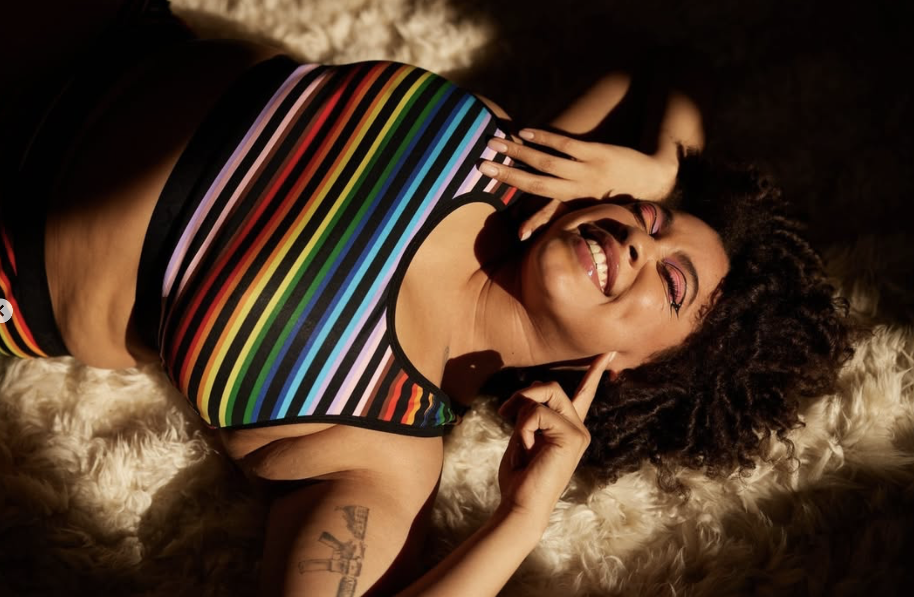 A woman lying on a fluffy surface, smiling joyfully, wearing a black top with rainbow-colored stripes, colorful makeup, and curly hair, with one hand near her face and the other resting on her chest.
