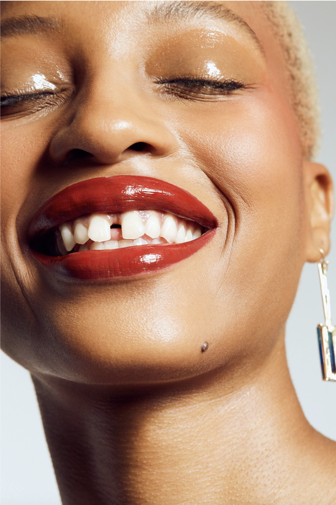 Close-up of a smiling woman with red lipstick, hoop earring, and light skin, showing her teeth, with closed eyes and a confident expression.