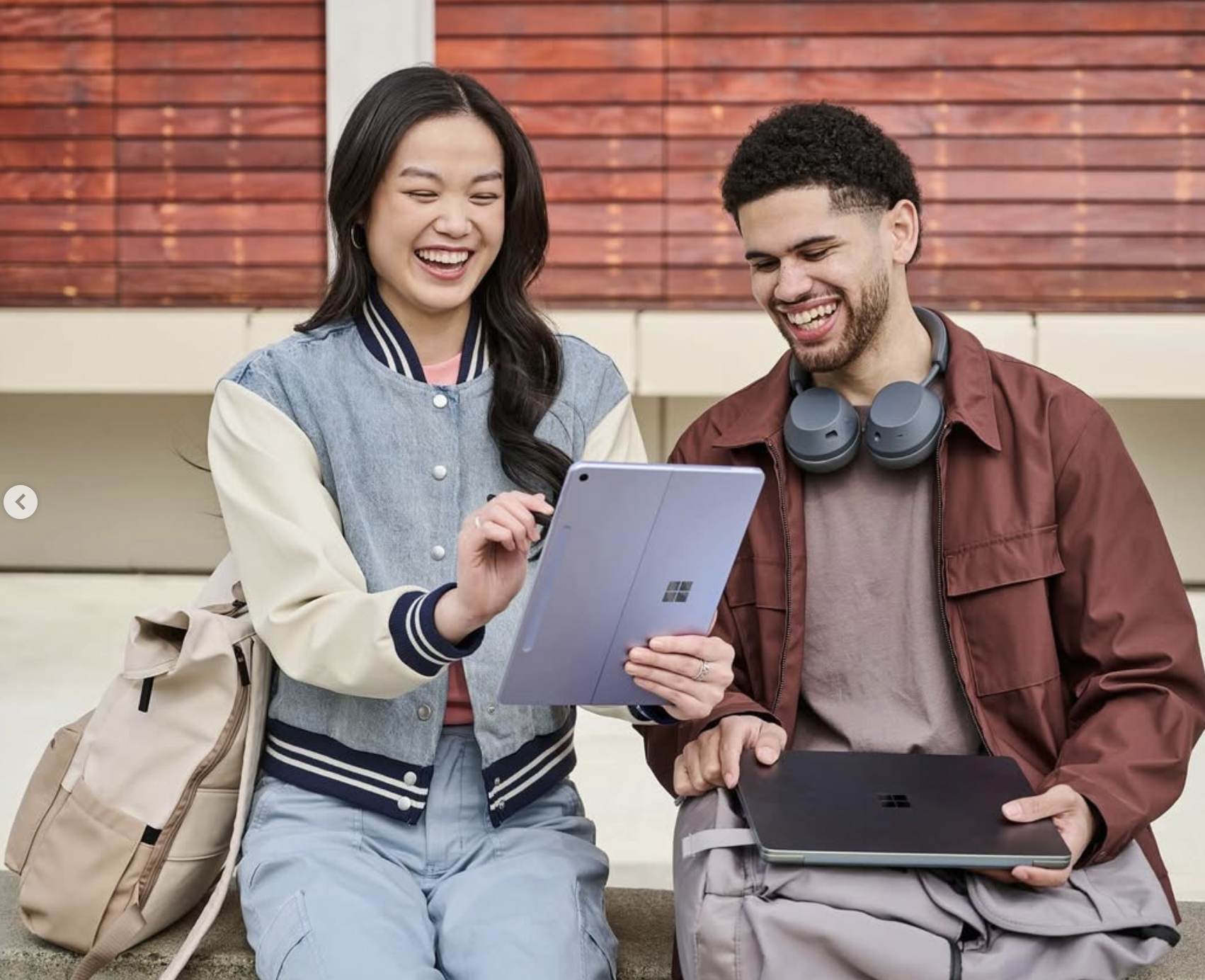 A young woman and man sitting outdoors, looking at a tablet and smiling, with a backpack and a laptop nearby.