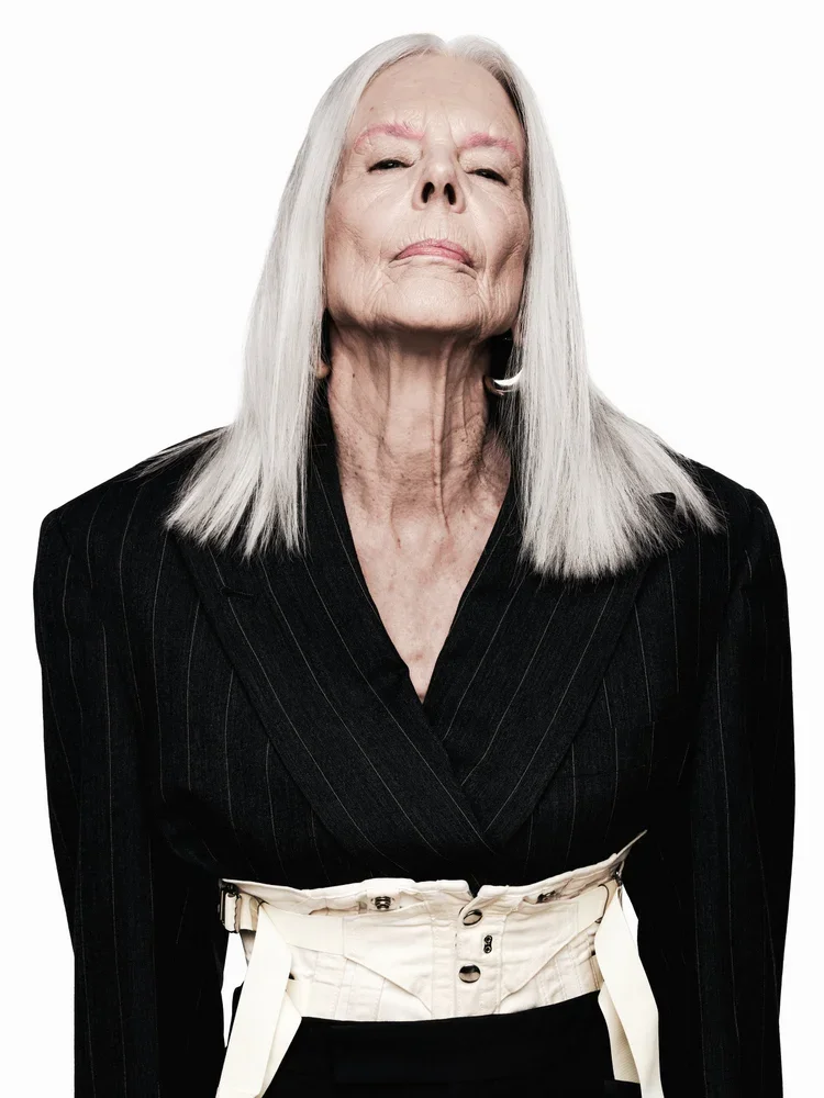 Portrait of an mature woman with long white hair, wearing a black pinstripe blazer and high-waisted beige pants, standing against a white background.