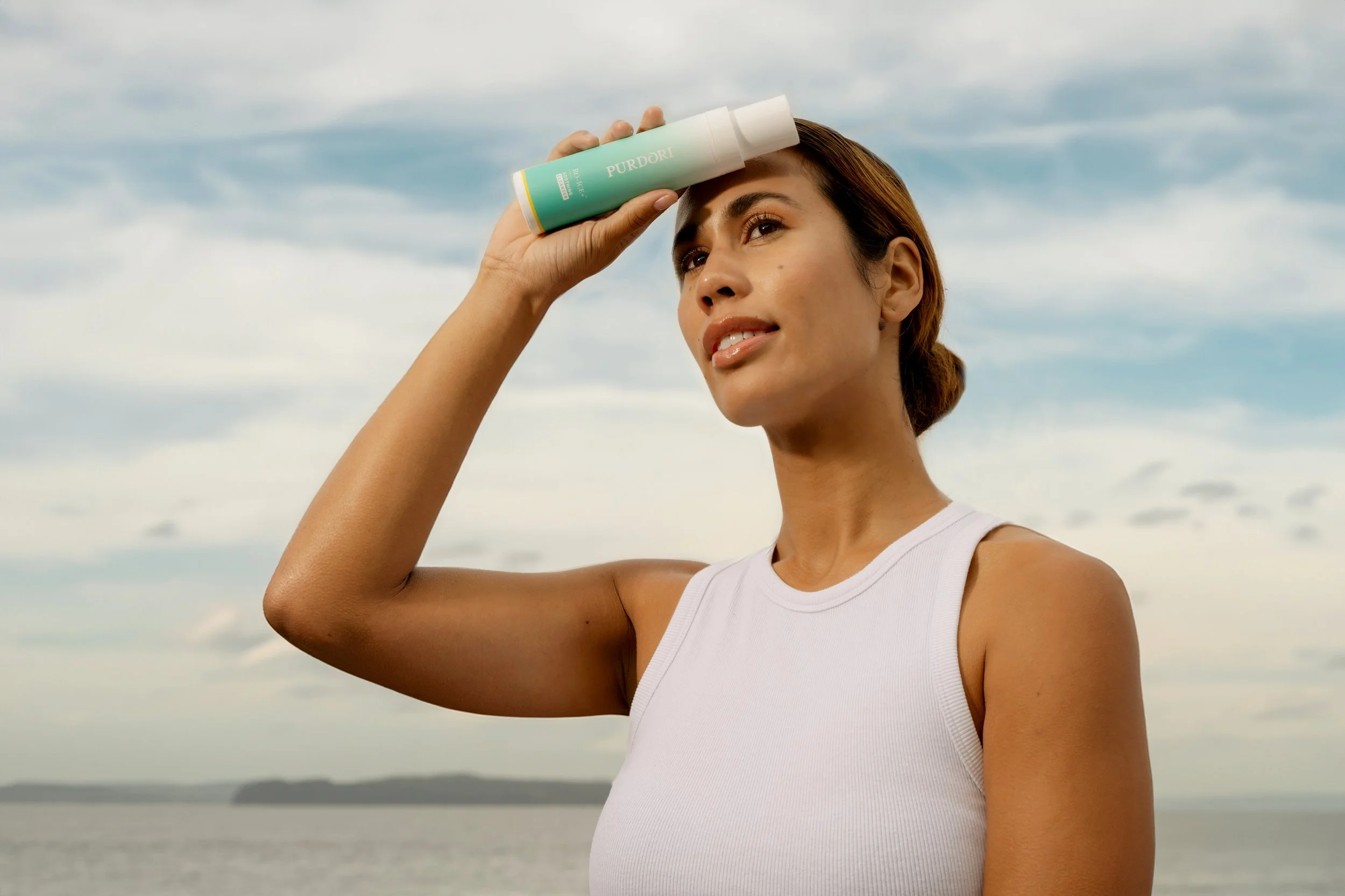 Woman with brown hair in a bun, wearing a white sleeveless top, holding a sunscreen bottle labeled 'Purdori' to her forehead outdoors by the water during cloudy weather.
