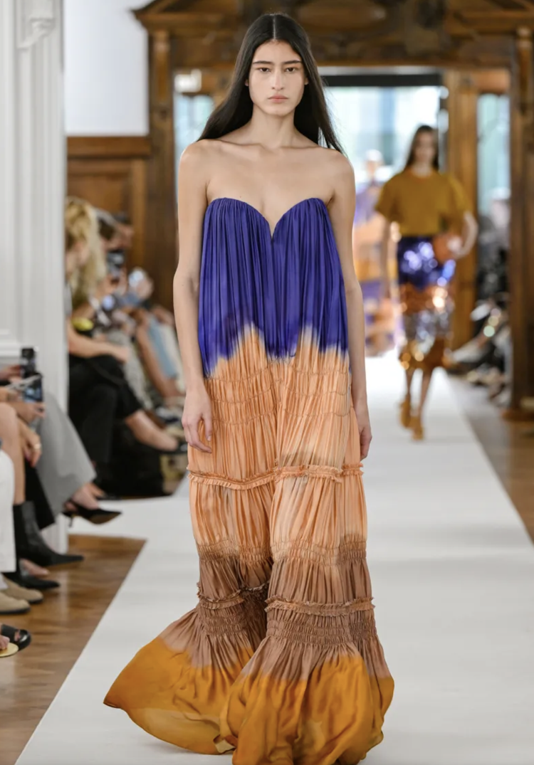 Model walking down runway in colorful, strapless, pleated maxi dress with blue, orange, and yellow tones, during a fashion show.