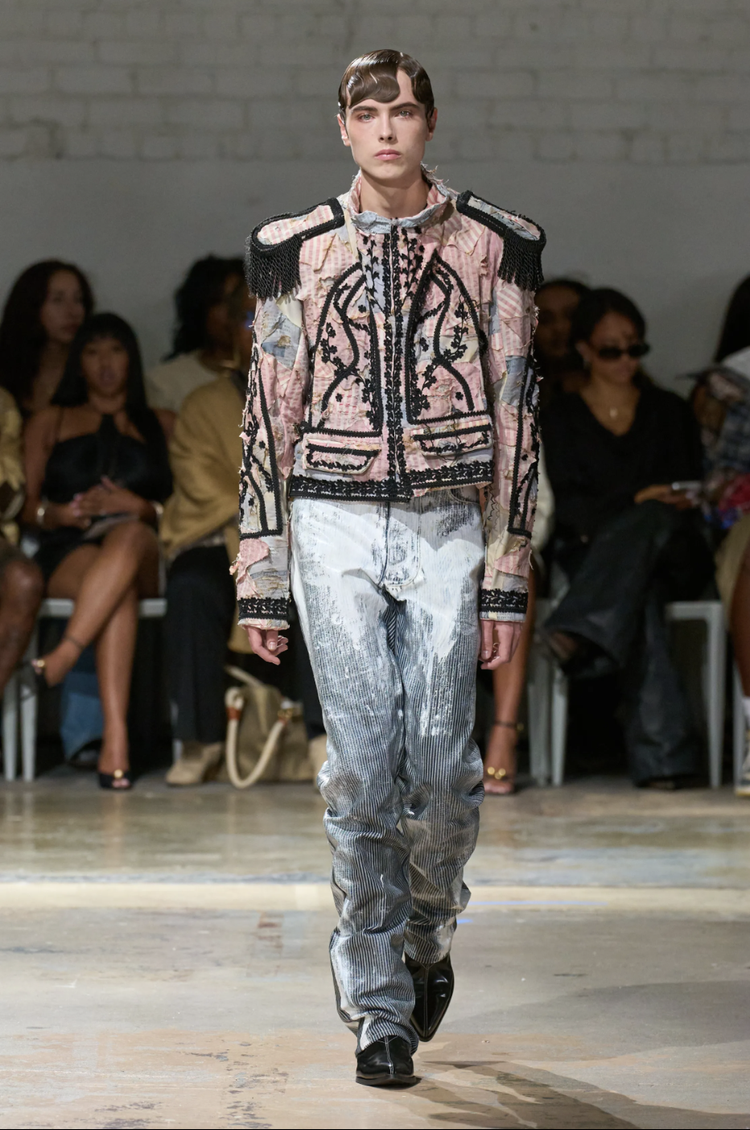 Fashion model walking on runway, wearing designer jacket with embroidery and shoulder epaulets, and metallic silver pants, audience seated in the background.