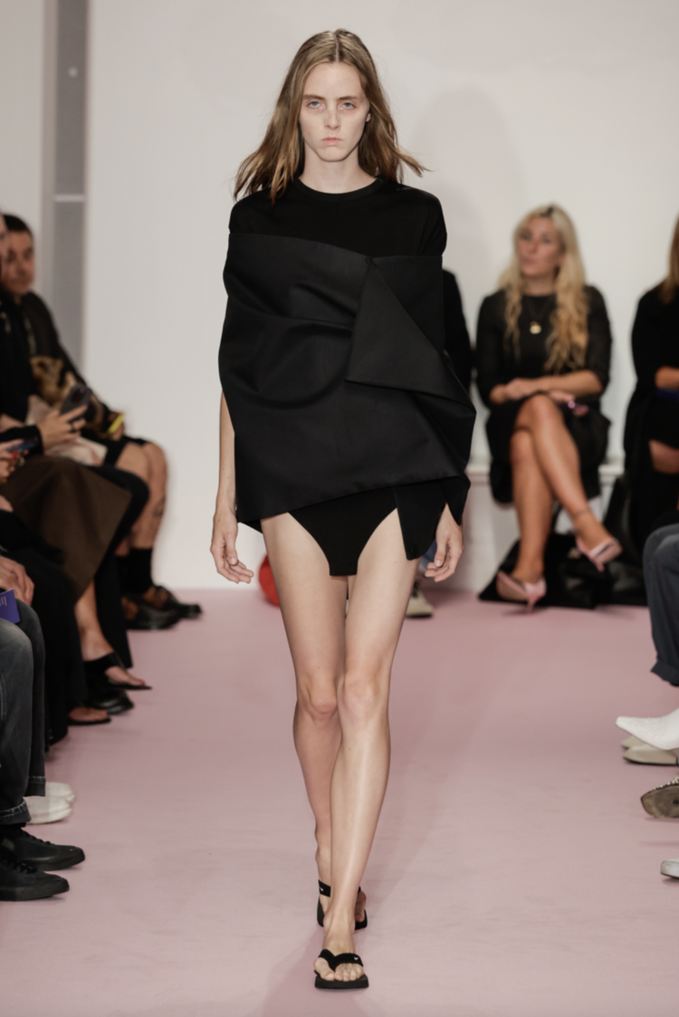 Model walking down runway during a fashion show, wearing an oversized black top with asymmetrical design, black high-heeled sandals, and black shorts, with an audience seated on both sides of the pink runway.