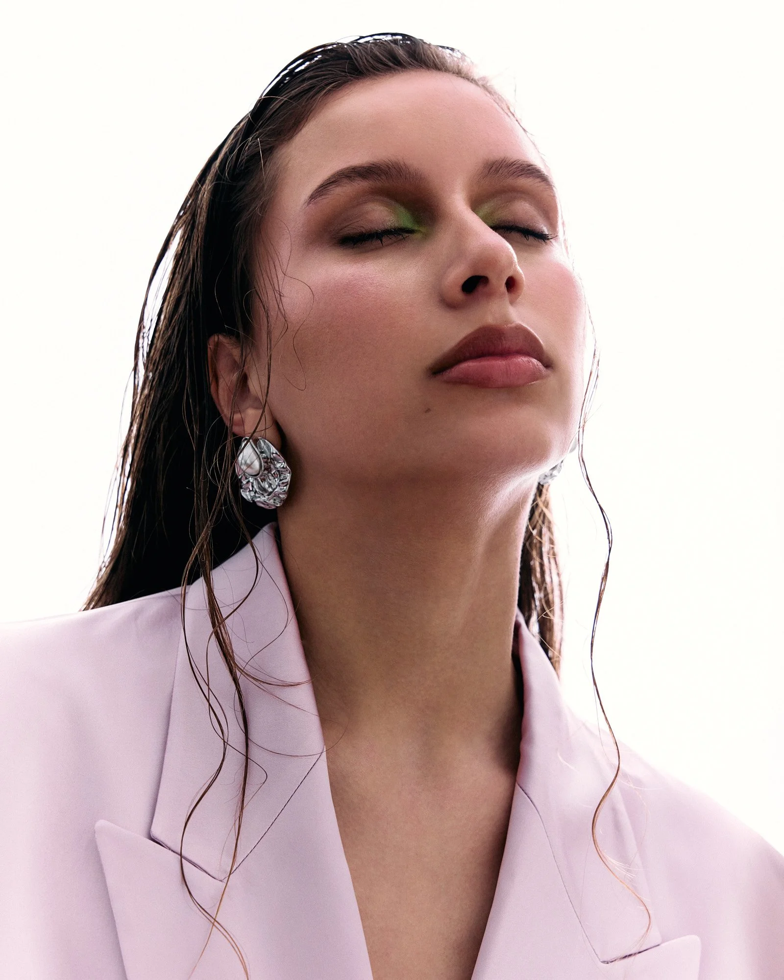 Close-up of a woman with wet hair, wearing a pink blazer and silver earrings, eyes closed, with a serene expression.