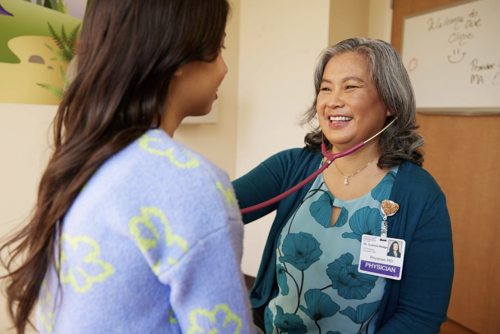A female doctor using a stethoscope to listen to a young woman's heartbeat in an examination room, both smiling.
