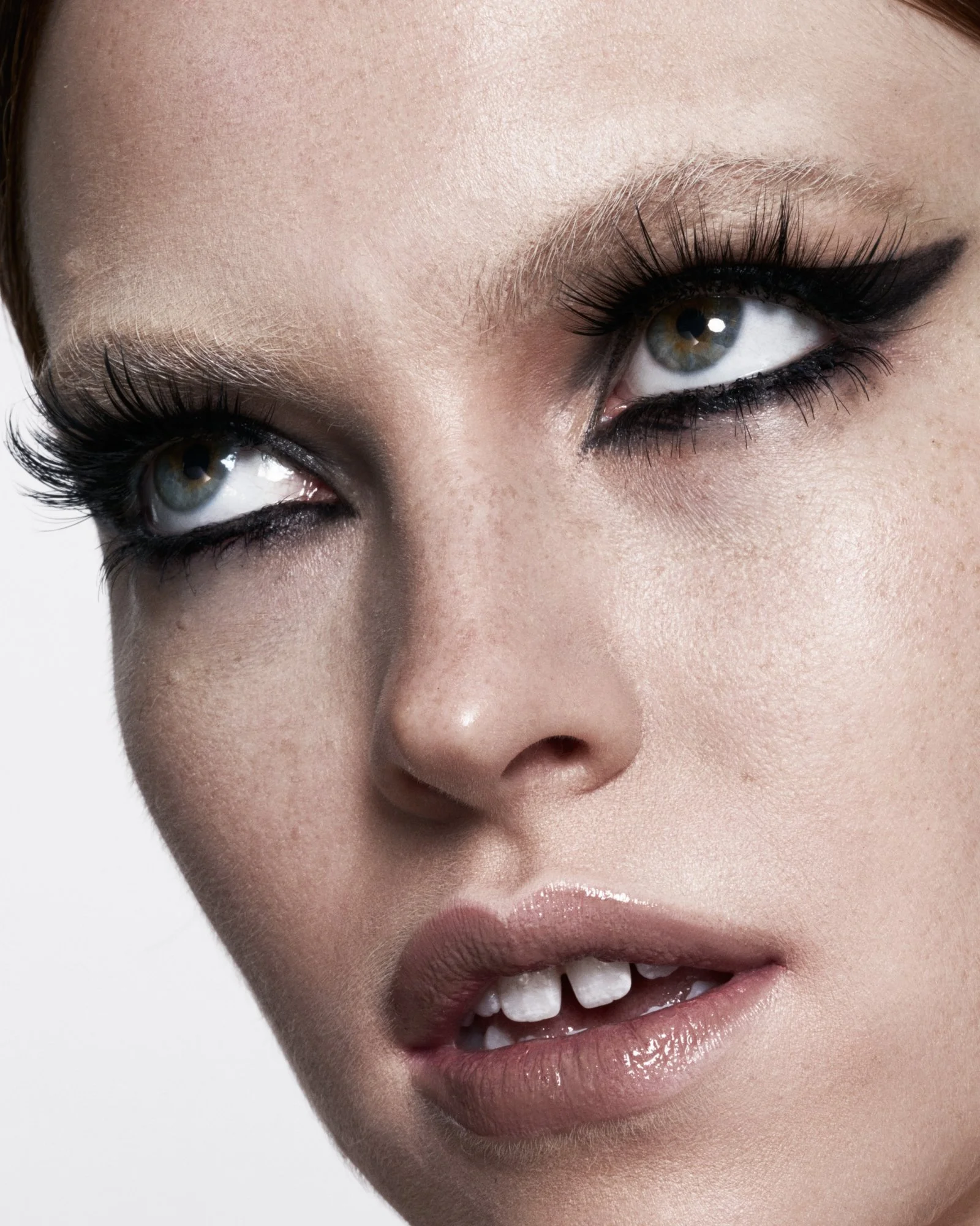 Close-up of a woman's face emphasizing her eyes with dramatic black eyeliner and long lashes, smooth skin, and slightly open lips showing teeth.