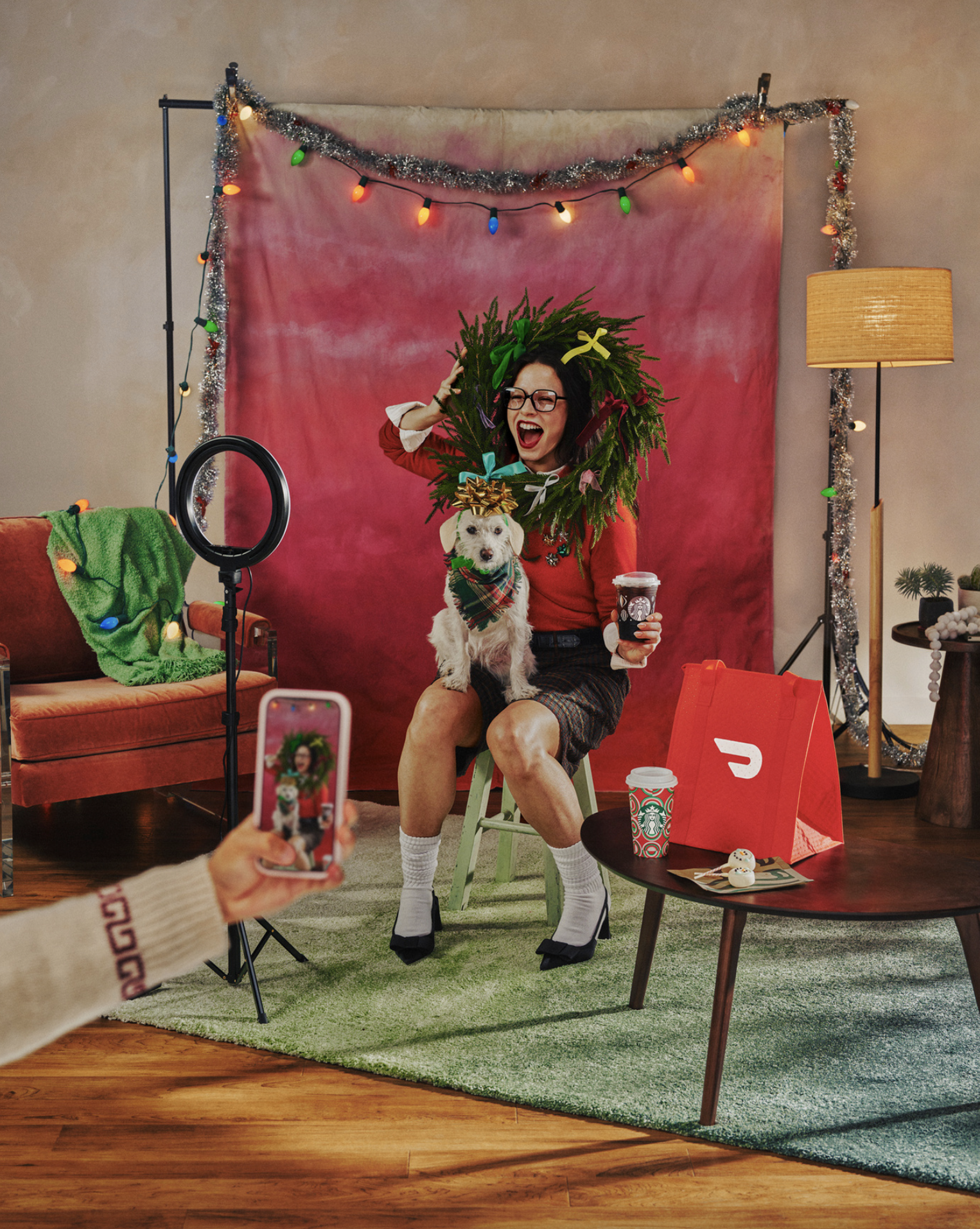 Woman with glasses, celebrating with a dog in a Christmas wreath, sitting on a stool in a festive room with decorations, while another person records her on a phone.