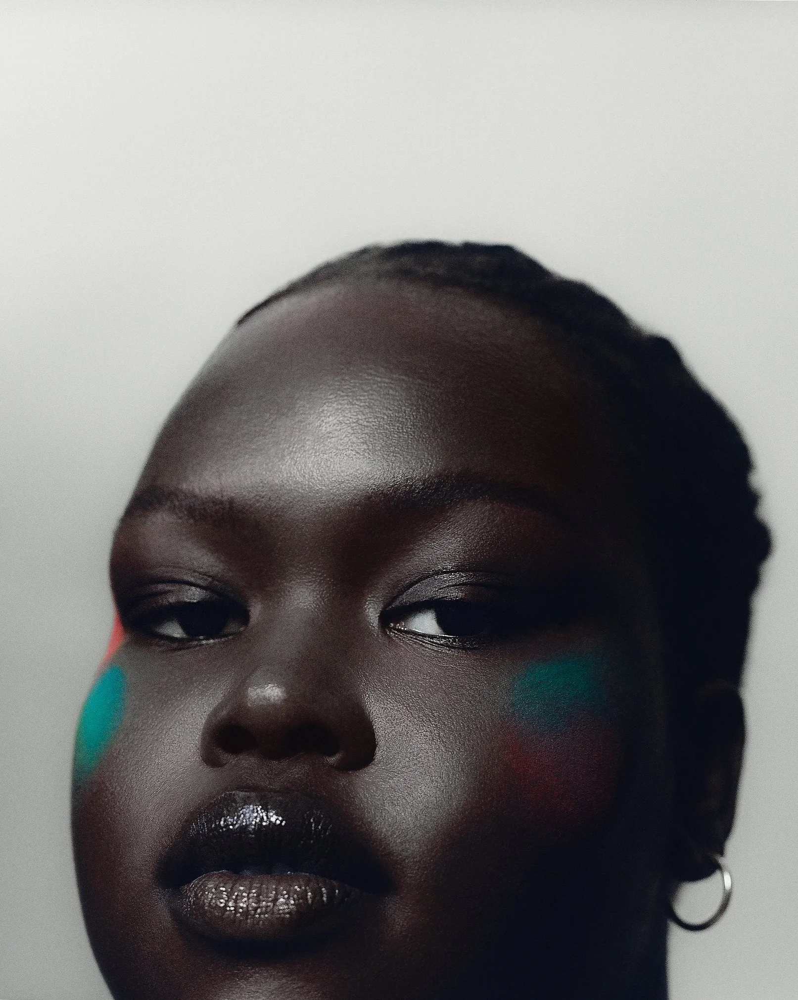 Close-up of a woman with dark skin, styled hair, bold black lipstick, eye makeup, with colorful face paint in teal, red, and orange on her cheeks, against a plain light background.