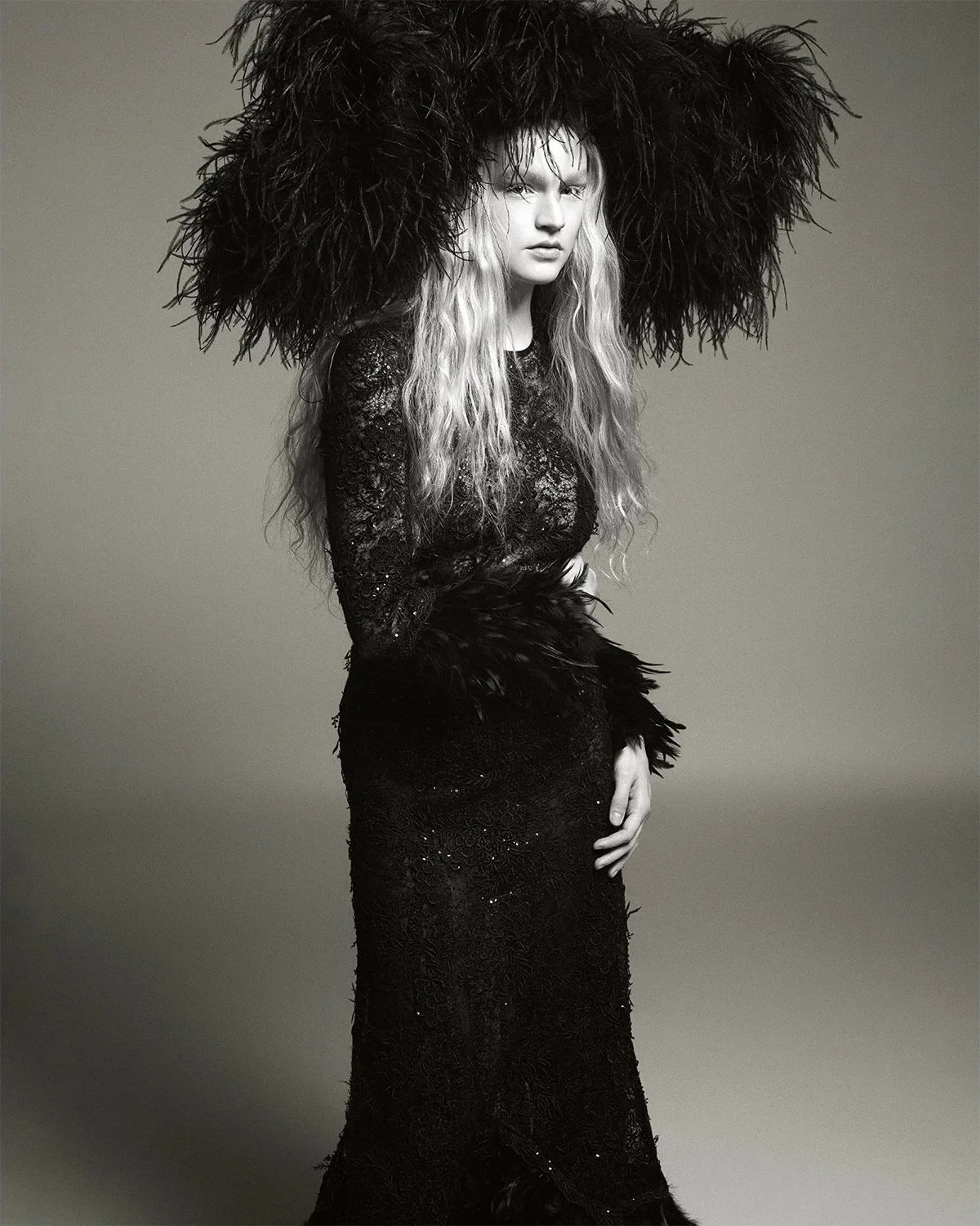 A woman with long curly blonde hair wearing a black lace dress with a large feathered hat.