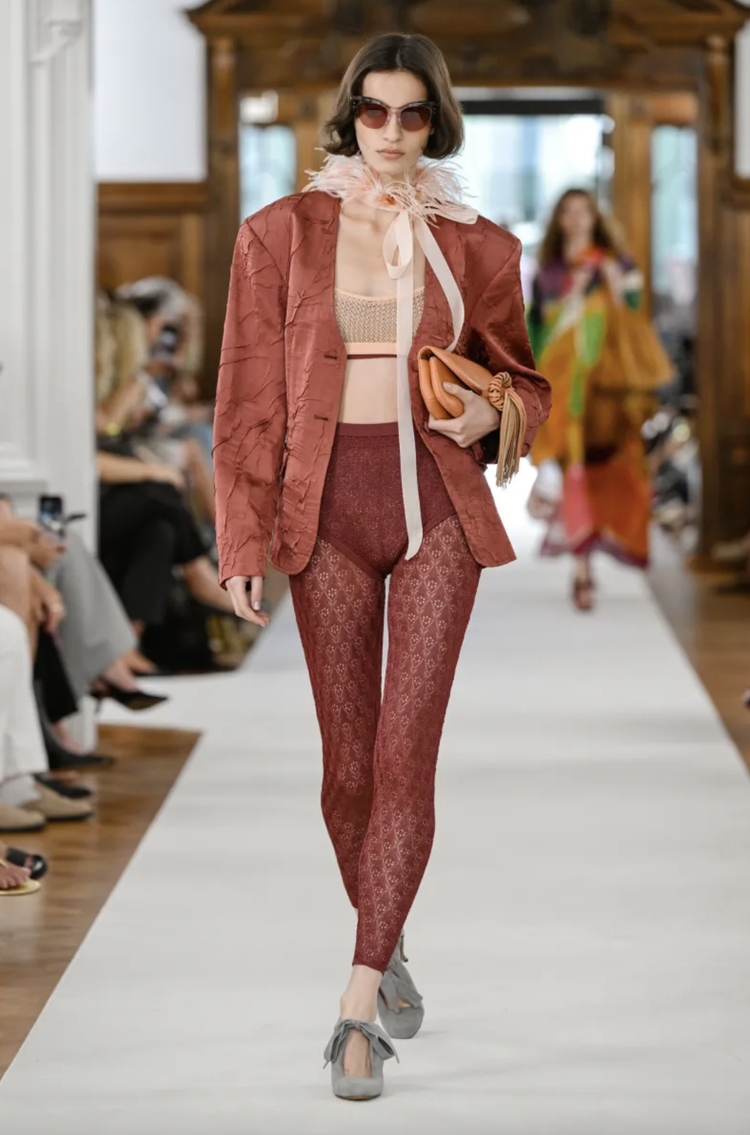Model walking on a runway wearing sunglasses, a pink feathered neck ribbon, a maroon textured blazer, a beige mesh top, maroon patterned pants, and gray shoes with bows, while holding an orange clutch in her left hand.