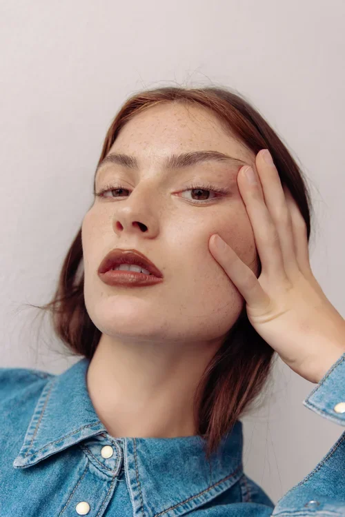 A young woman with red hair and freckles, wearing a denim jacket, posing with her hand on the side of her face against a plain background.