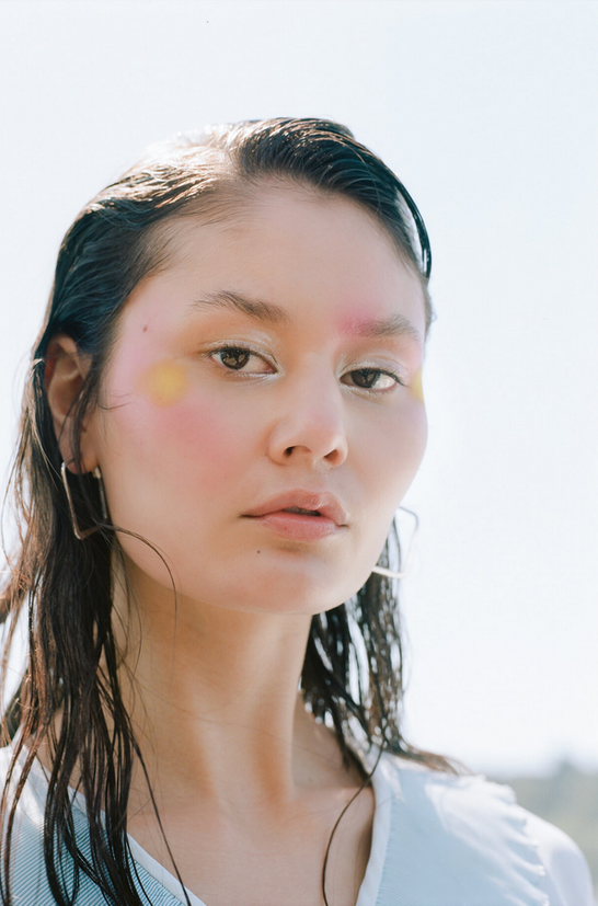A young woman with wet hair and natural makeup standing outdoors against a bright sky.