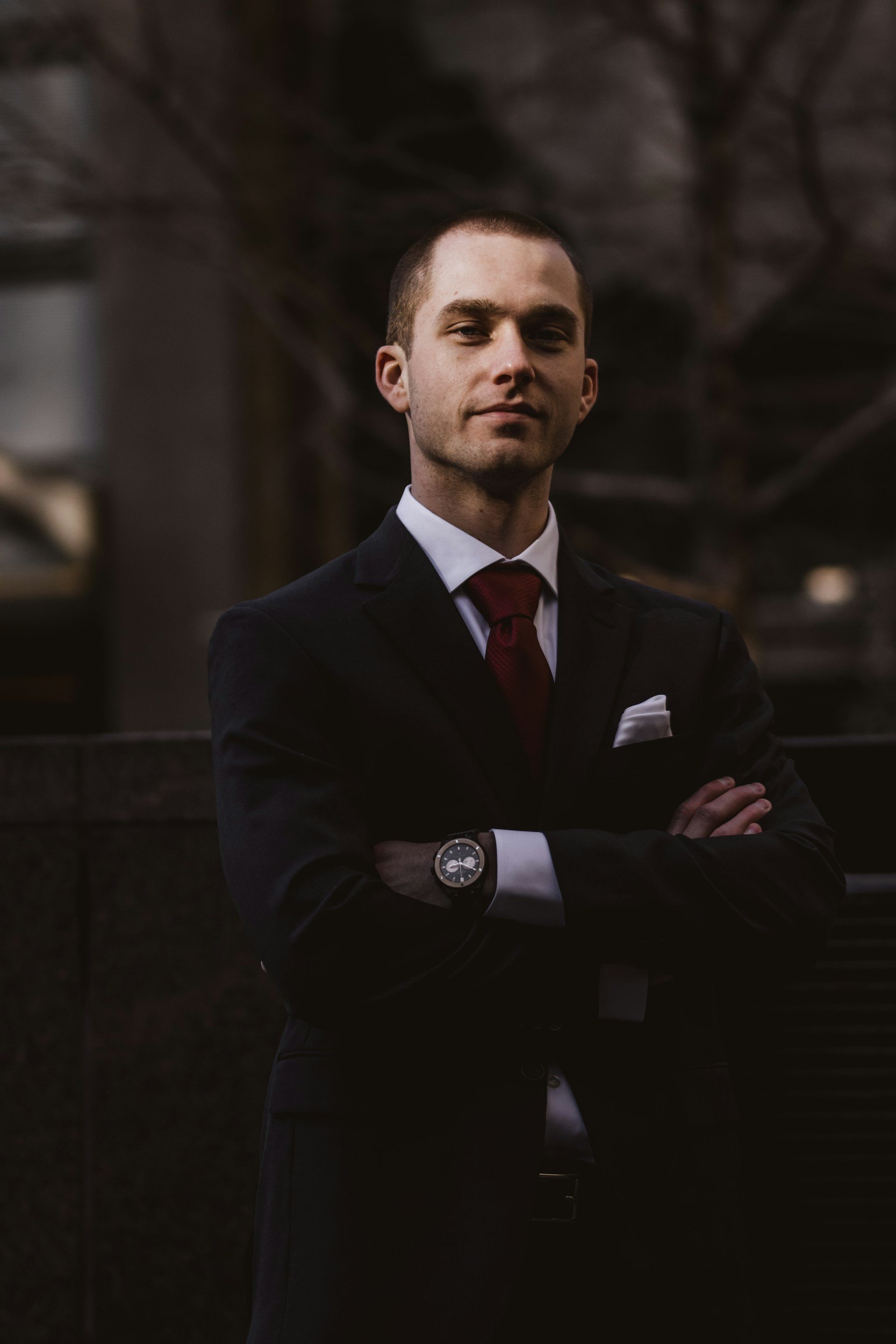 A young man in a black suit, white shirt, red tie, and a watch on his left wrist, standing outdoors with arms crossed, facing the camera with a confident expression.