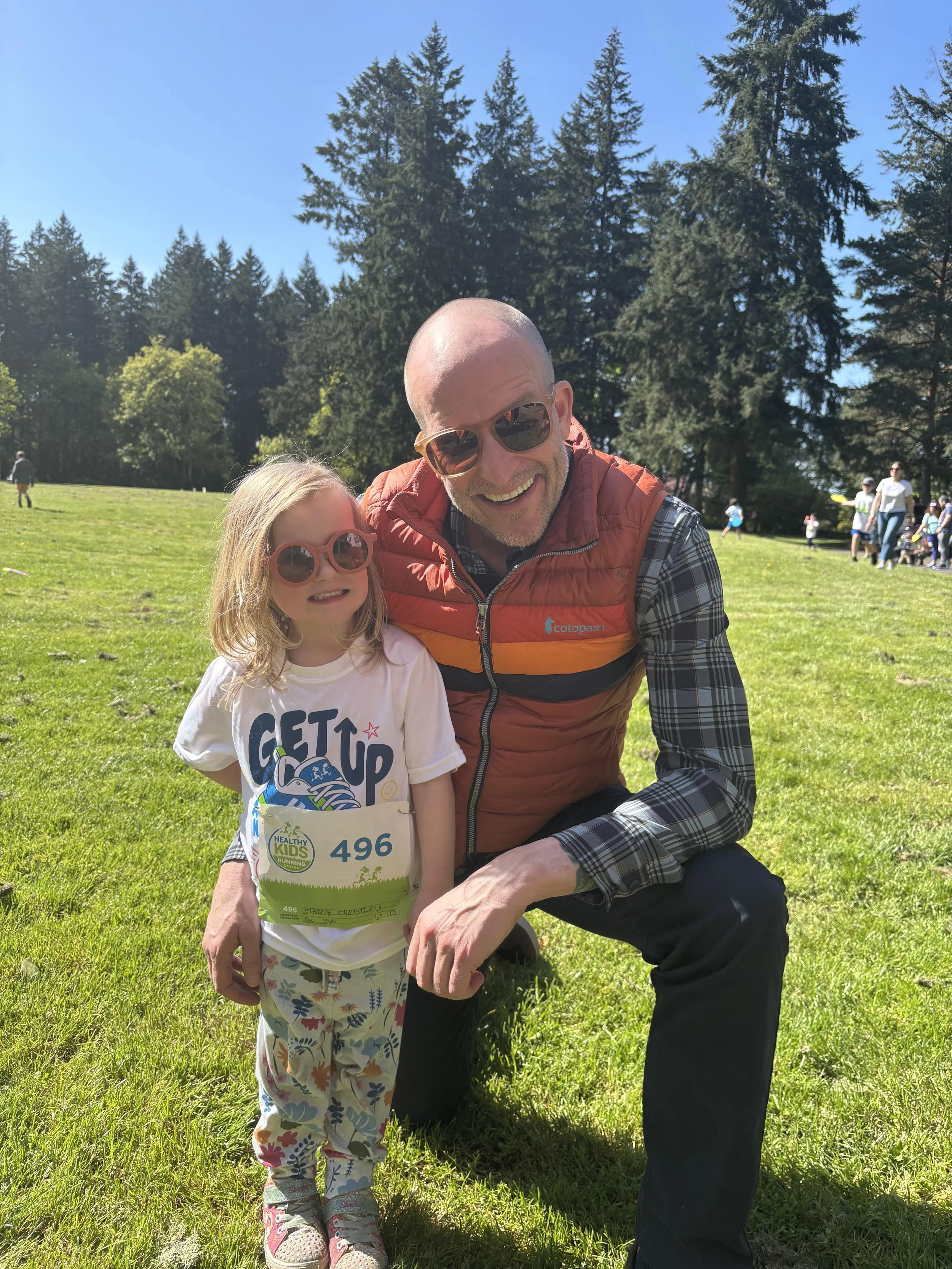 Ben and his daughter outdoors on a sunny day, both wearing sunglasses. The girl wears a white T-shirt with 'Get Up' and a race bib number 496, along with patterned pants.