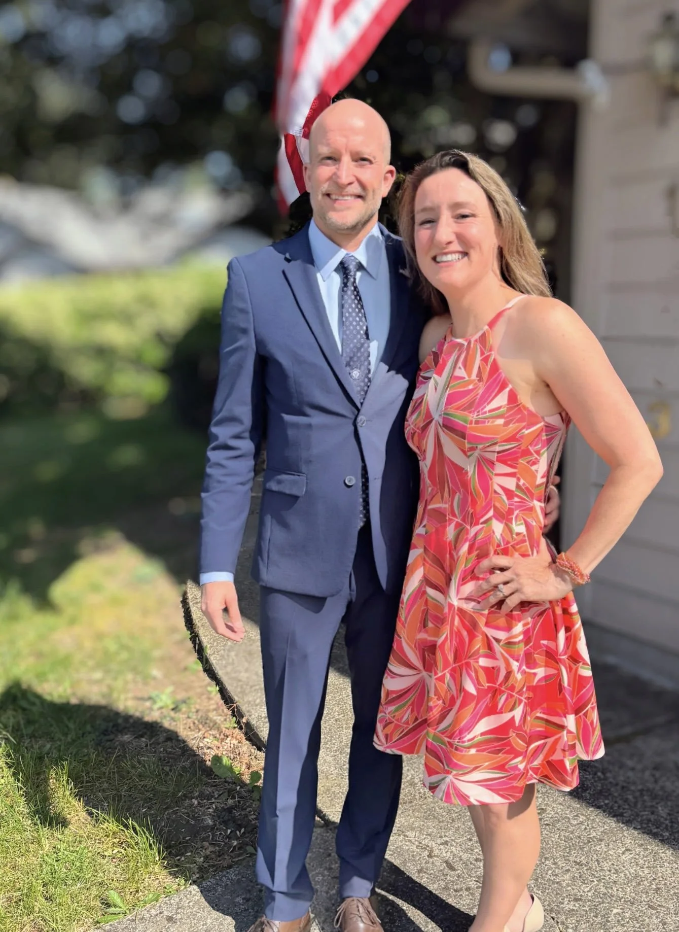 A man in a blue suit and a woman in a colorful sleeveless dress standing outdoors in front of an American flag, smiling for a photo.