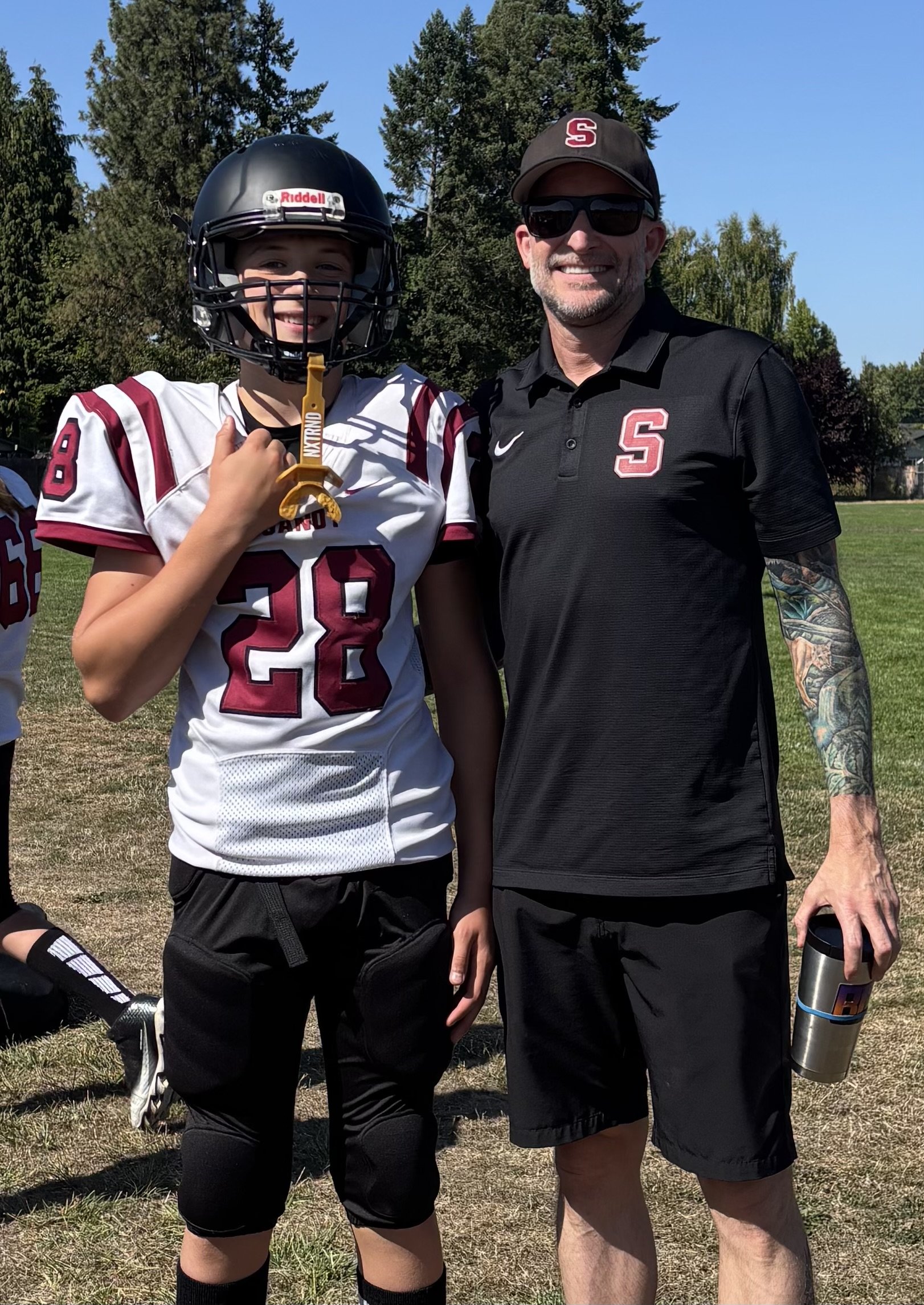 A young football player in uniform with a helmet, standing next to a man in a black sports shirt and sunglasses, both smiling outdoors on a grassy field with trees in the background.