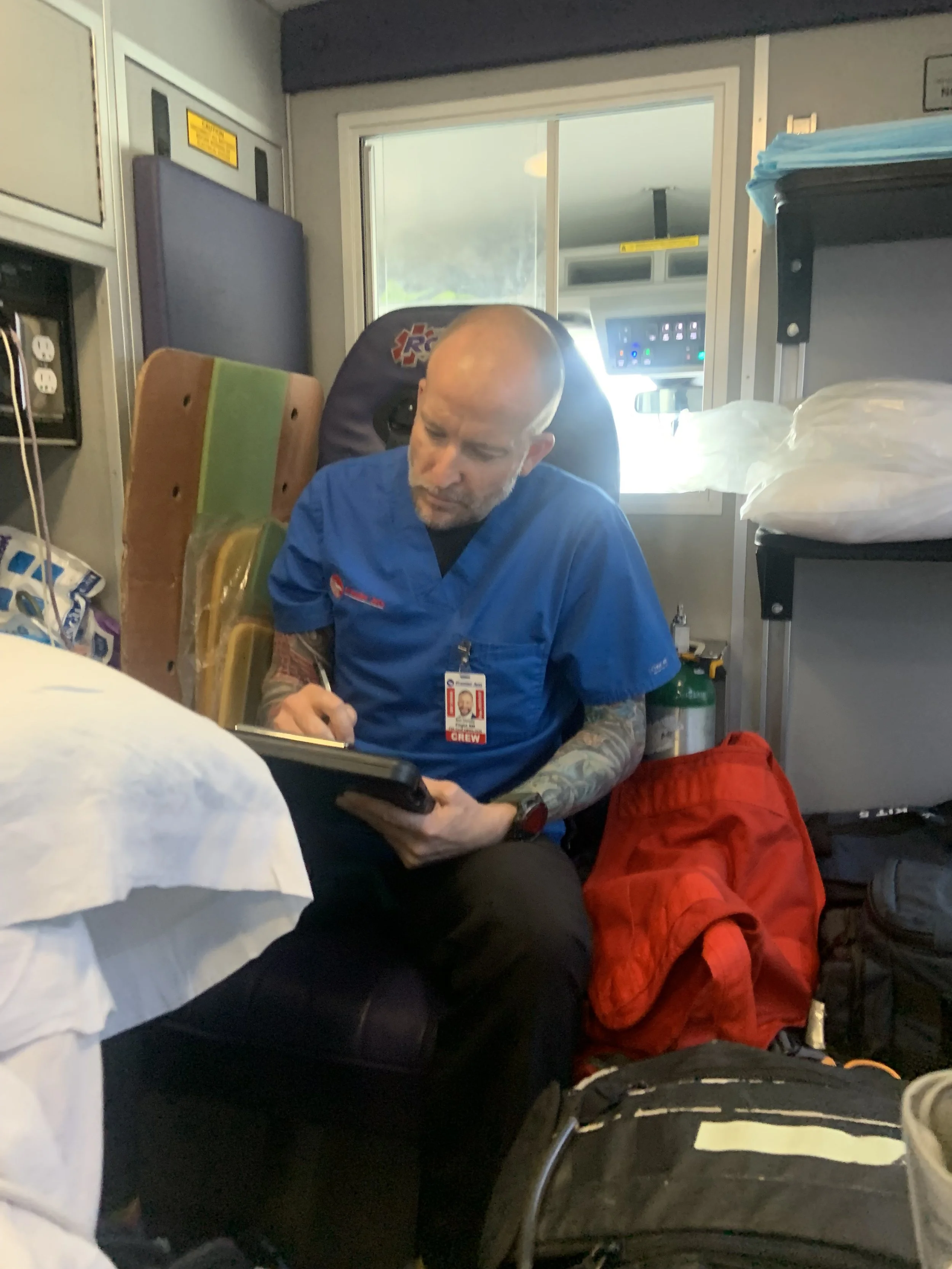 A male paramedic wearing a blue uniform and an ID badge, sitting in an ambulance, using a tablet, surrounded by medical supplies and equipment.