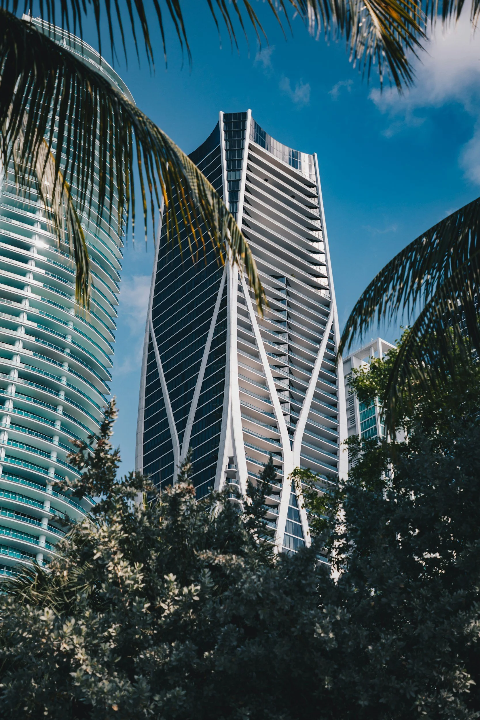 Tall modern skyscraper in a city, surrounded by palm trees and greenery, under a blue sky.